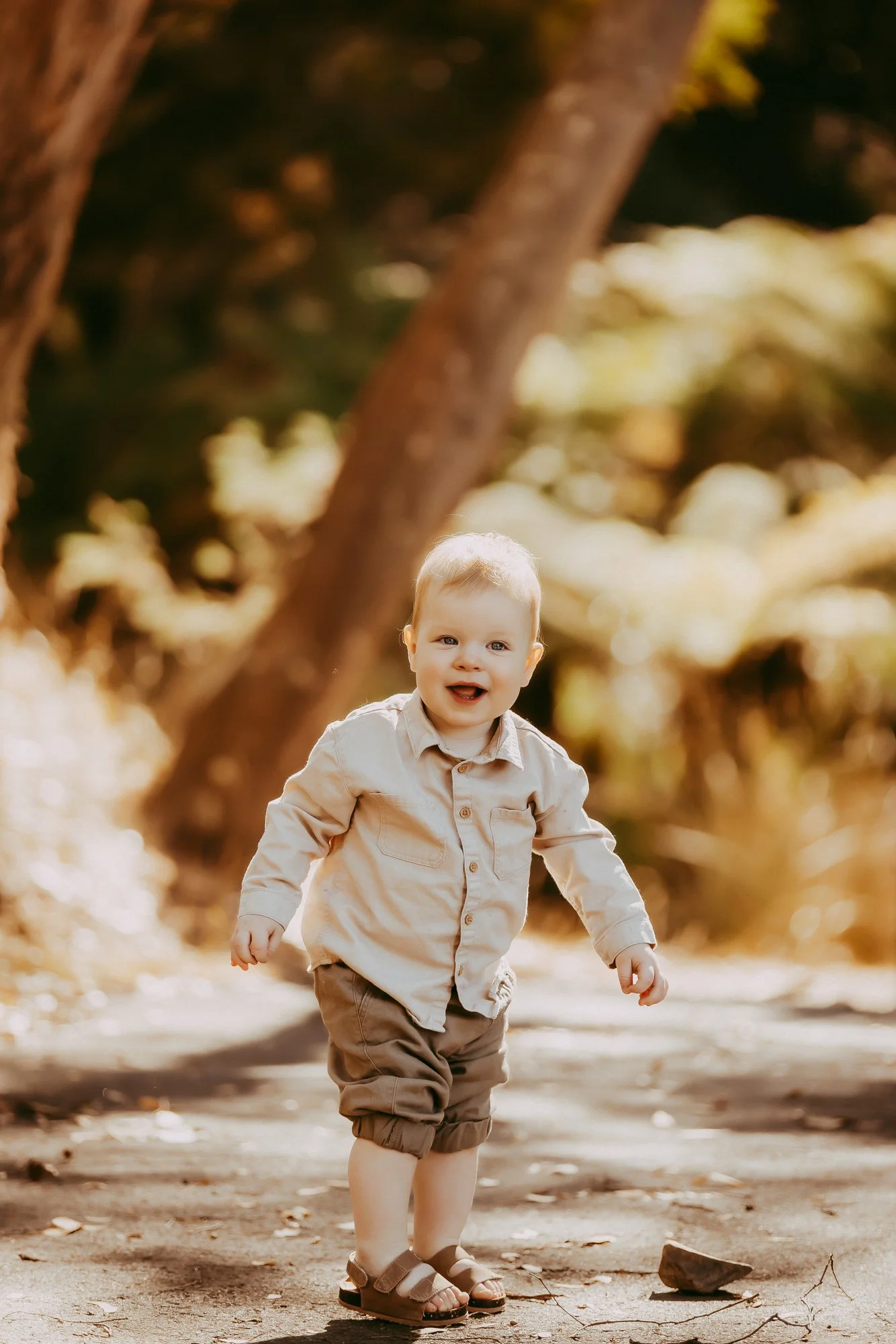 A young child with blonde hair smiling and walking outdoors on a sunny day, surrounded by trees and fallen leaves. Affordable family photographer in Greater Hobart area.
