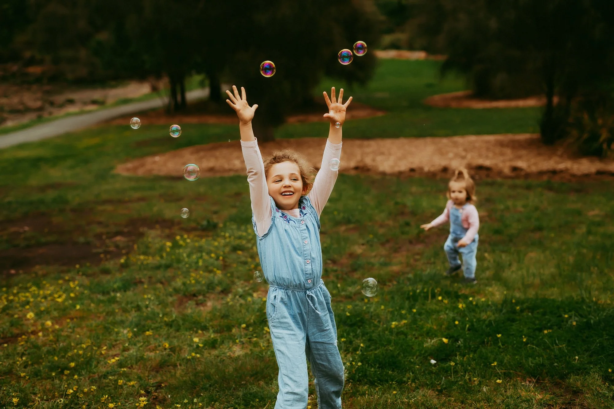 Two young girls playing outside with soap bubbles in a park. The girl in the foreground is smiling with her arms raised, while the girl in the background is walking towards her. The park has green grass, trees, and a winding path.