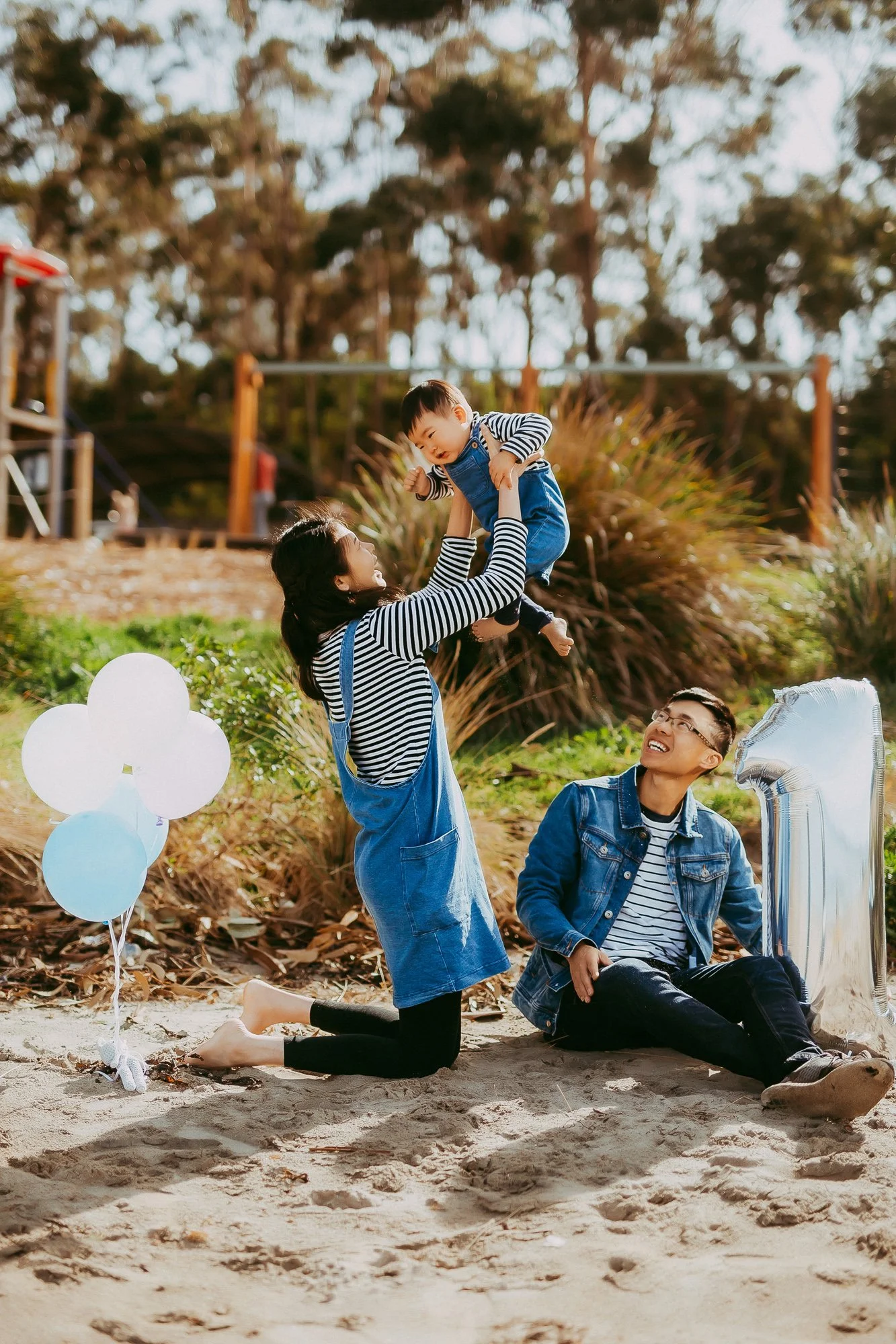 Family celebrating birthday on the beach with balloons, mother lifting child, father sitting with big silver number 1 balloon.