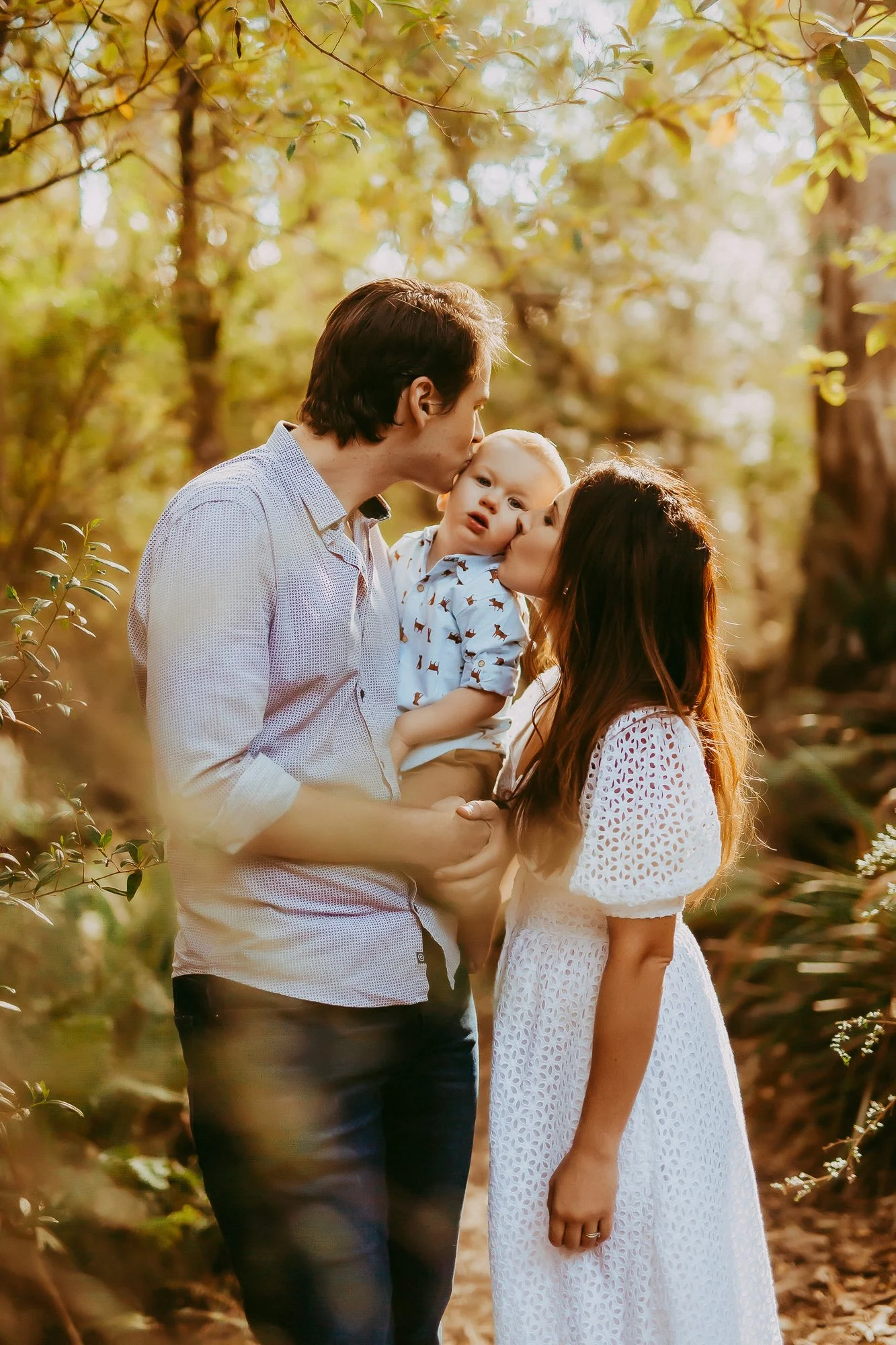 A family of three sharing a kiss outdoors in a forest during golden hour. The father is holding their young child, and the mother is kissing the child's cheek.