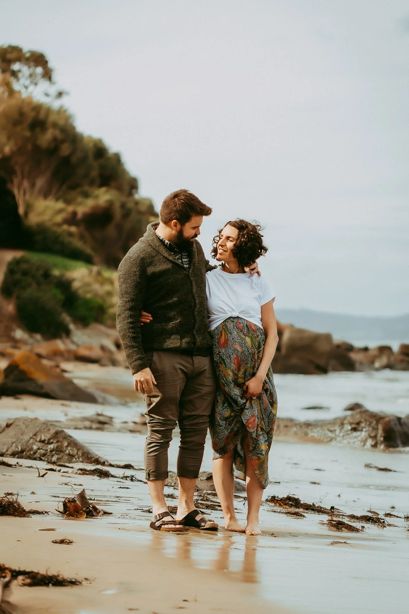 A couple walking barefoot on a beach, smiling and looking at each other, with waves and rocky shoreline in the background. Greater Hobart family photographer.