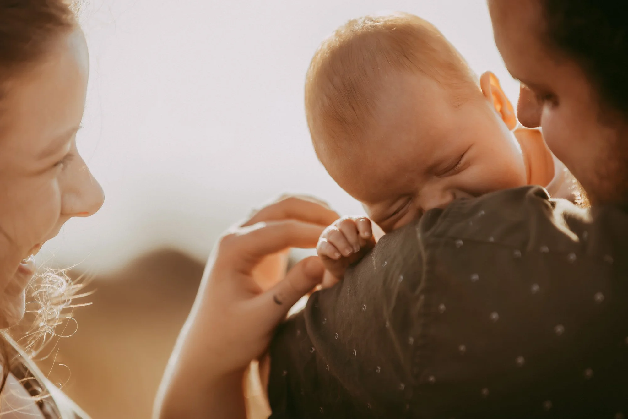 A man holding a sleeping baby close to his face while a woman looks on and gently touches the baby's hand in a bright, softly lit setting. Golden hour family photosession on the beach.