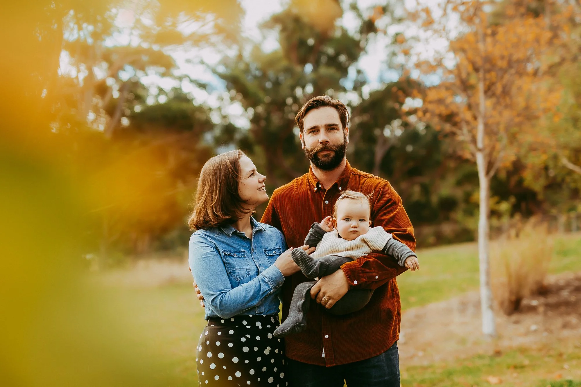 A family of three standing outdoors in a park during autumn. The father holds a baby, while the mother looks at the baby. The background features trees with fall foliage.