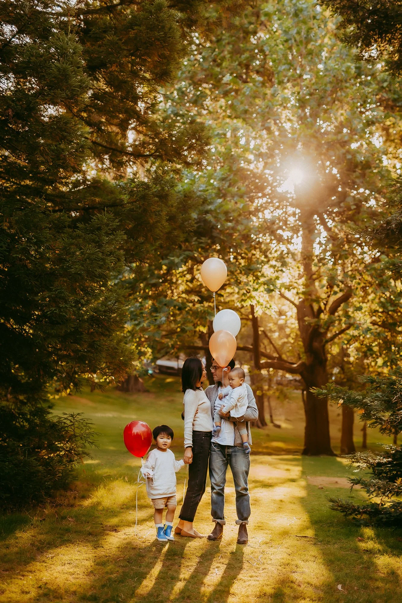 A family of four enjoying a sunny afternoon in a park, surrounded by trees and holding balloons. Budget friendly family photoshoot.