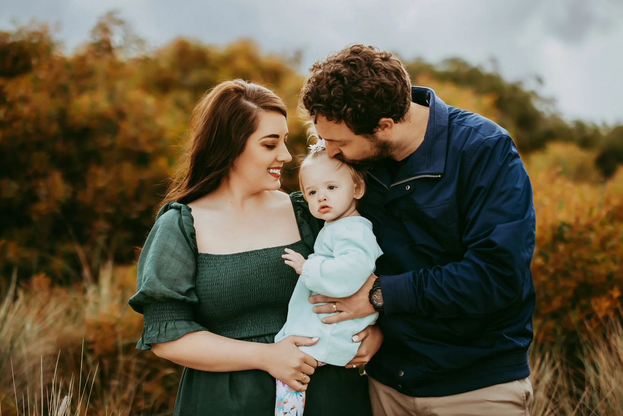 A family of three, a woman, a man, and a small girl, are outdoors with autumn-colored trees in the background. The woman is smiling at the girl, while the man leans in to kiss the girl on the head, creating a warm, loving scene.