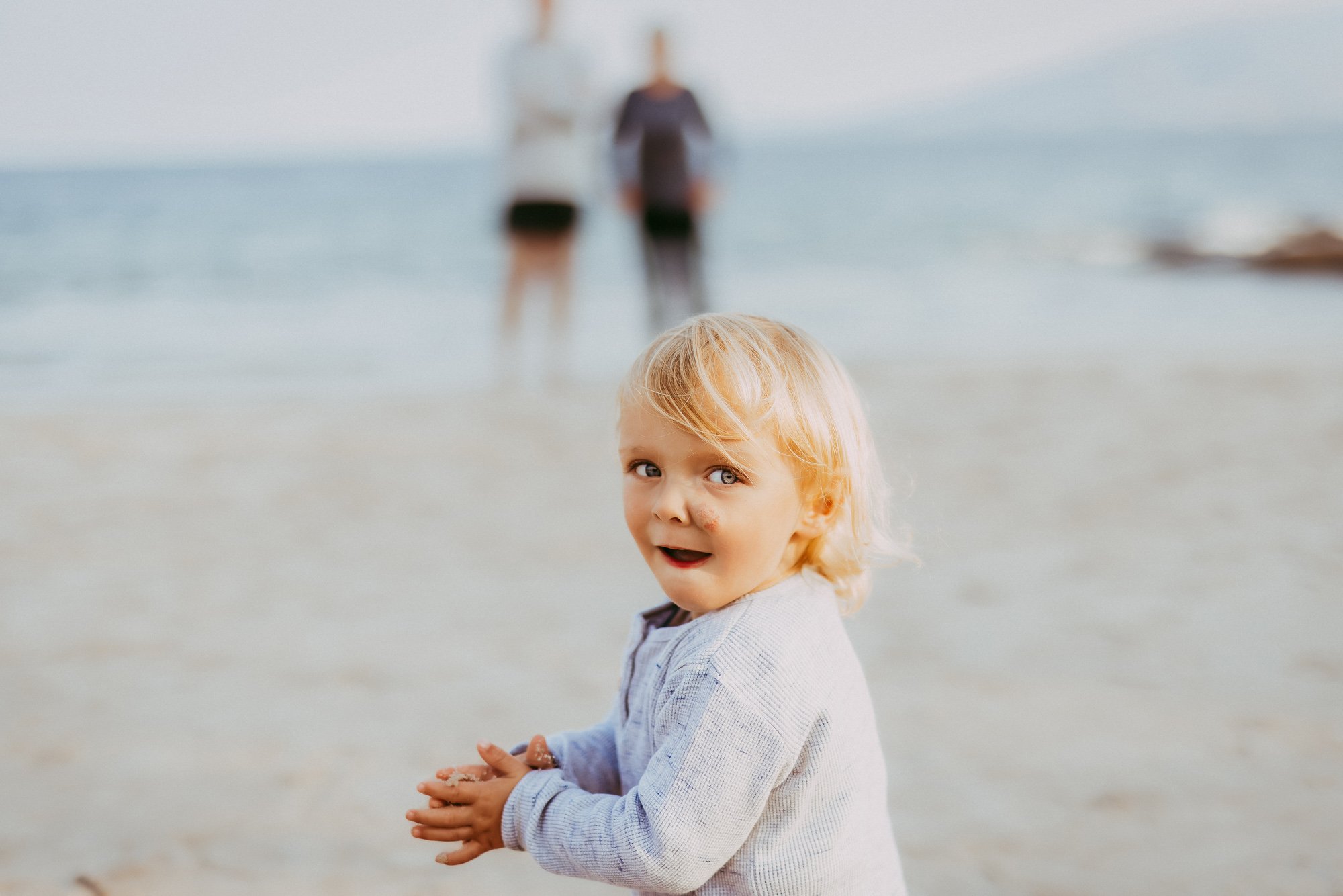A young blonde child with sandy hands standing on the beach, smiling at the camera, with a blurred couple in the background near the water.
