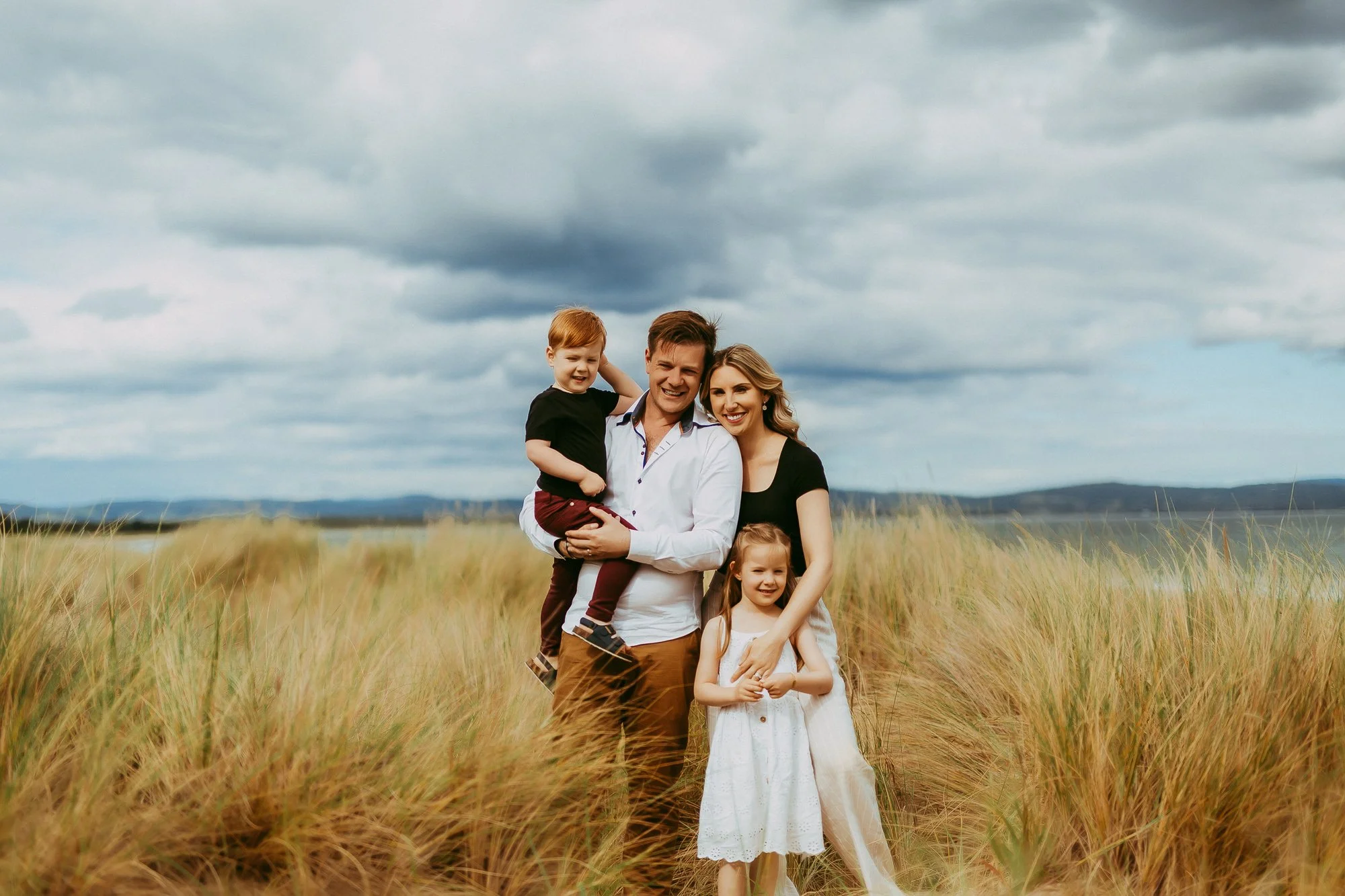 A family of four standing in a field of tall grass with dark, cloudy skies above.