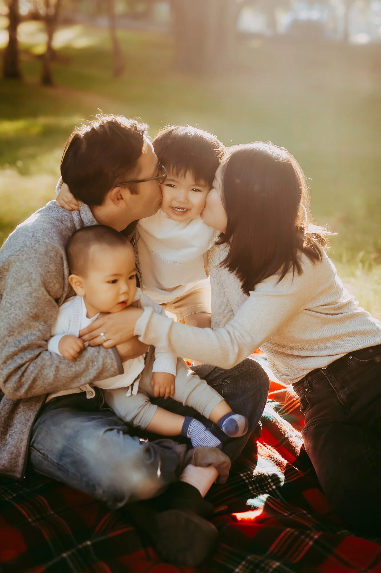 A family of four sits on a plaid blanket outdoors near a pond, with sunlight filtering through trees, as the parents kiss their kids on the cheeks. Affordable family photoshoot.