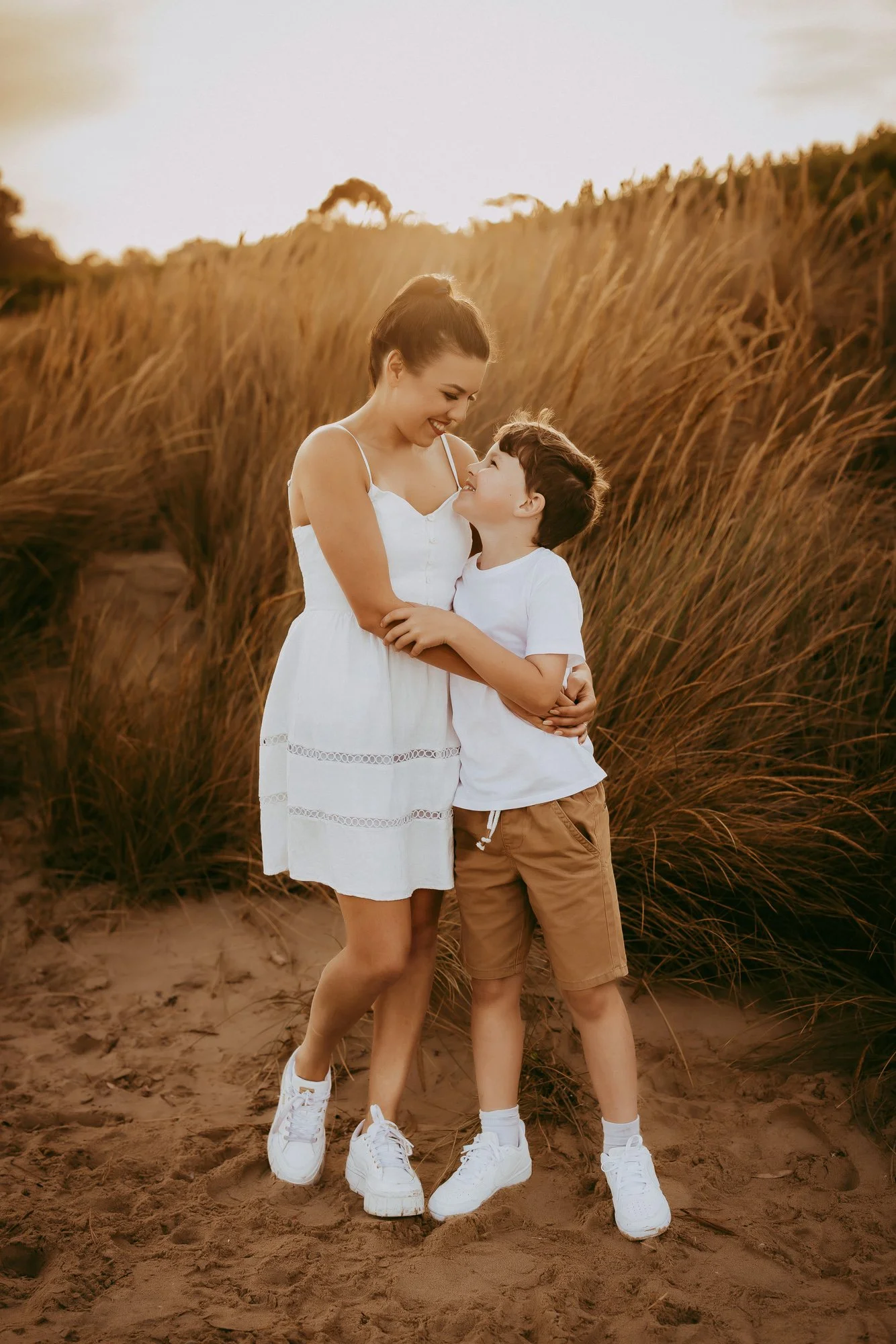 A woman and young boy sharing a happy moment outdoors in a field of tall grasses at sunset, smiling and embracing.