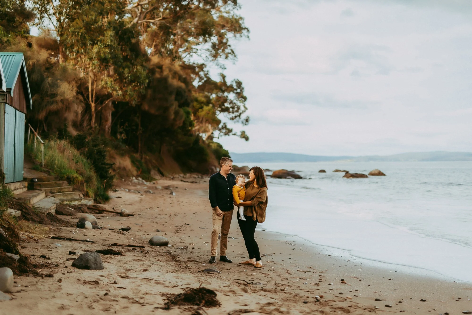 A family of three standing on the beach near the shoreline, with trees and rocks in the background, during a cloudy day.