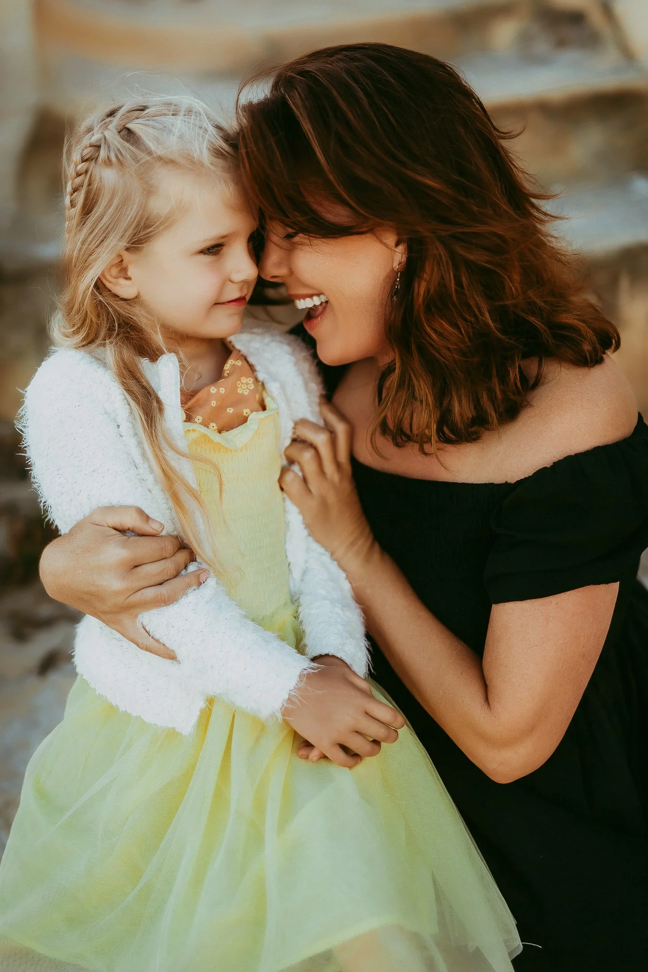 A woman and a young girl with blond hair sharing a close, affectionate moment, touching foreheads and smiling, outdoors with a blurred background. Unposed Hobart family photography