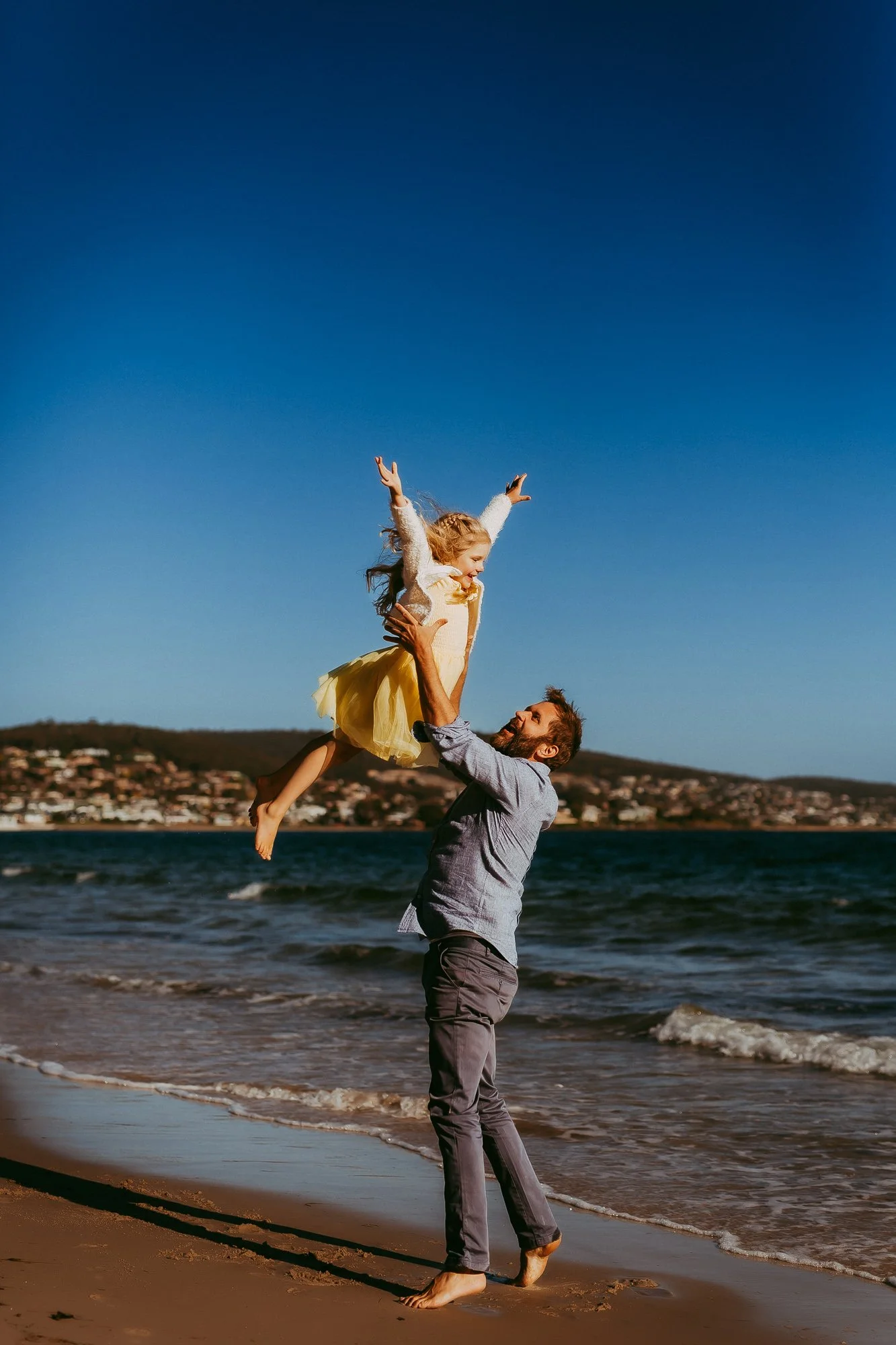 Man lifting girl in yellow skirt and white sweater on the beach with ocean and hills in the background. Greater Hobart family photosession with relaxed approach.