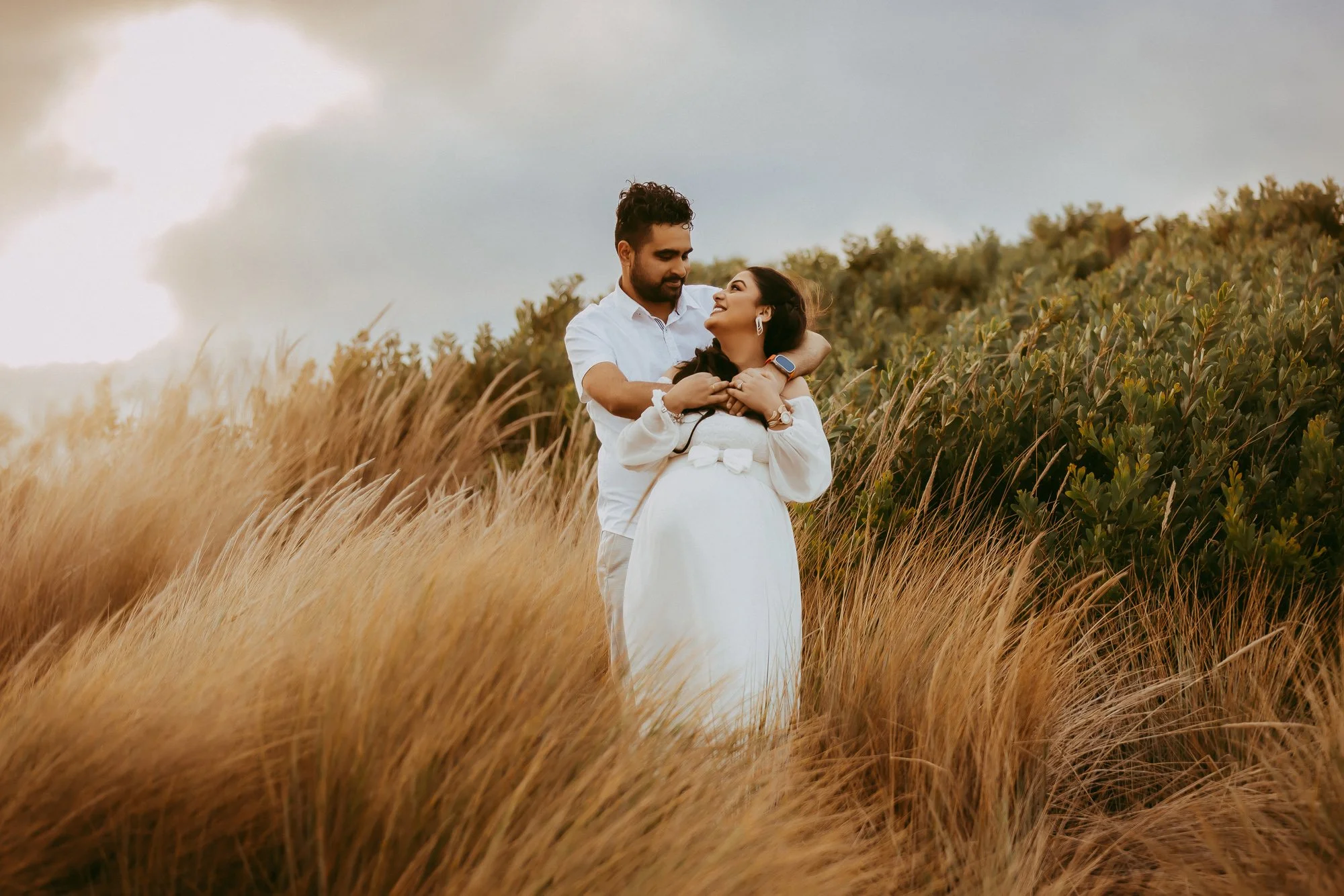 A man and woman embrace in a field of tall, golden grass, with green shrubbery in the background and a partly cloudy sky above.