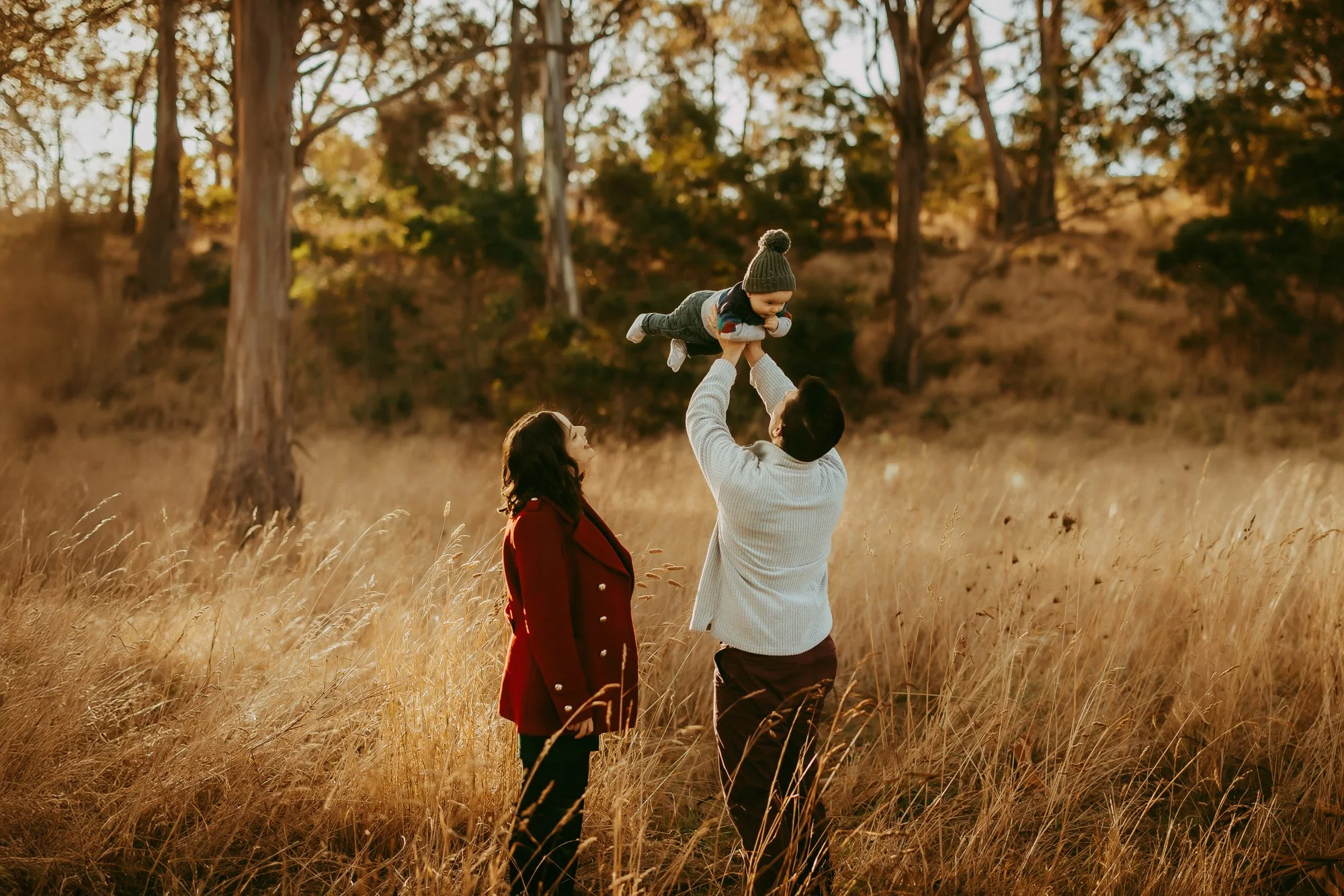 A man lifting a toddler in the air while a woman looks on in a field of tall grass during sunset with trees in the background. Golden hoer Hobart Family photoshoot by Ulla.
