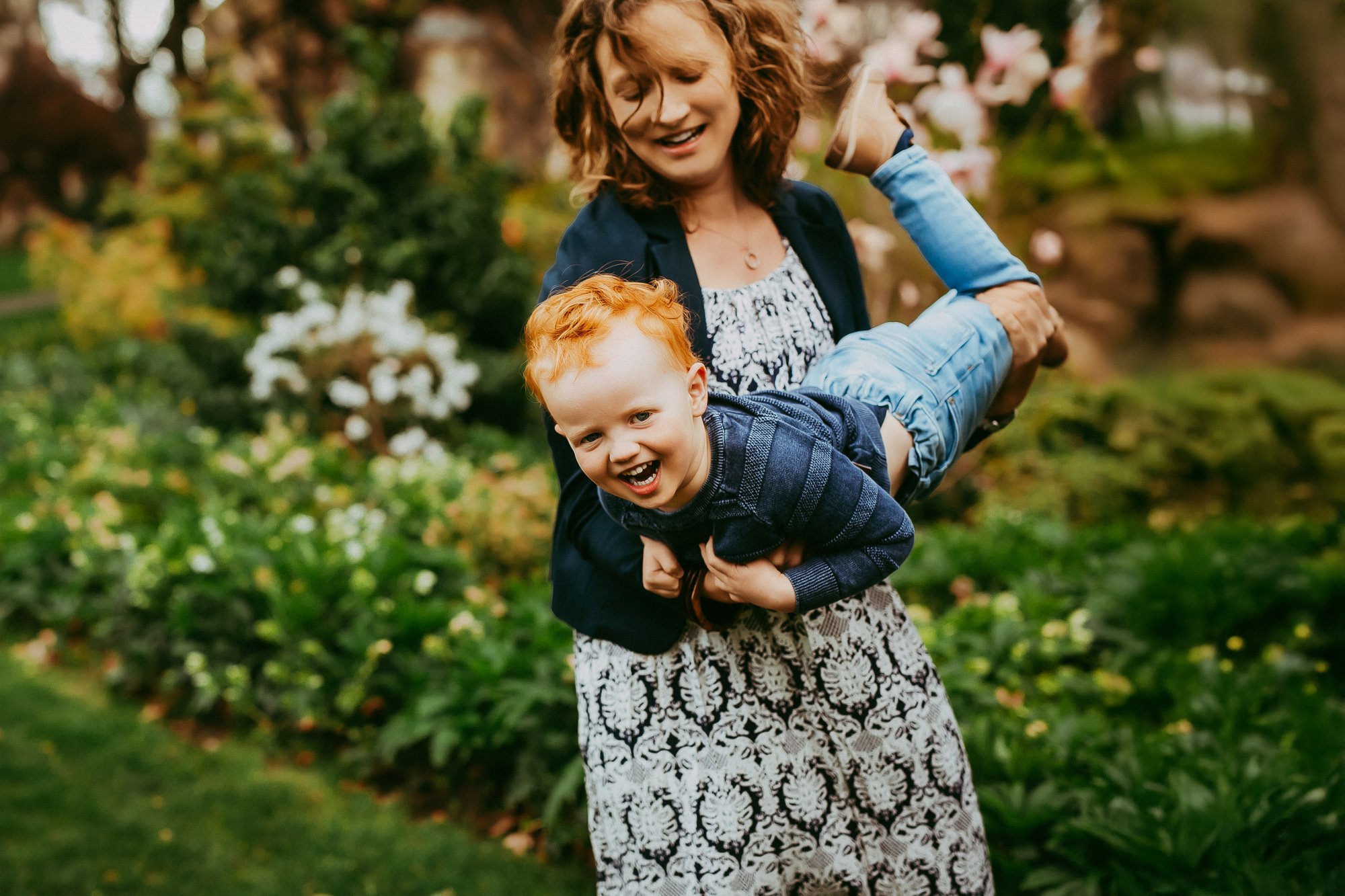 Woman smiling and holding a happy curly-haired boy horizontally in a garden with flowers and greenery. Family photosession by Ulla in Hobart area.