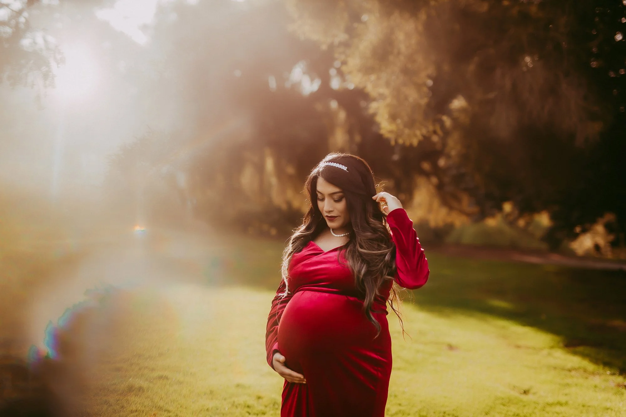 Pregnant woman in a red dress standing outdoors in a park during sunset or sunrise, gently holding her belly with her right hand and touching her hair with her left hand.