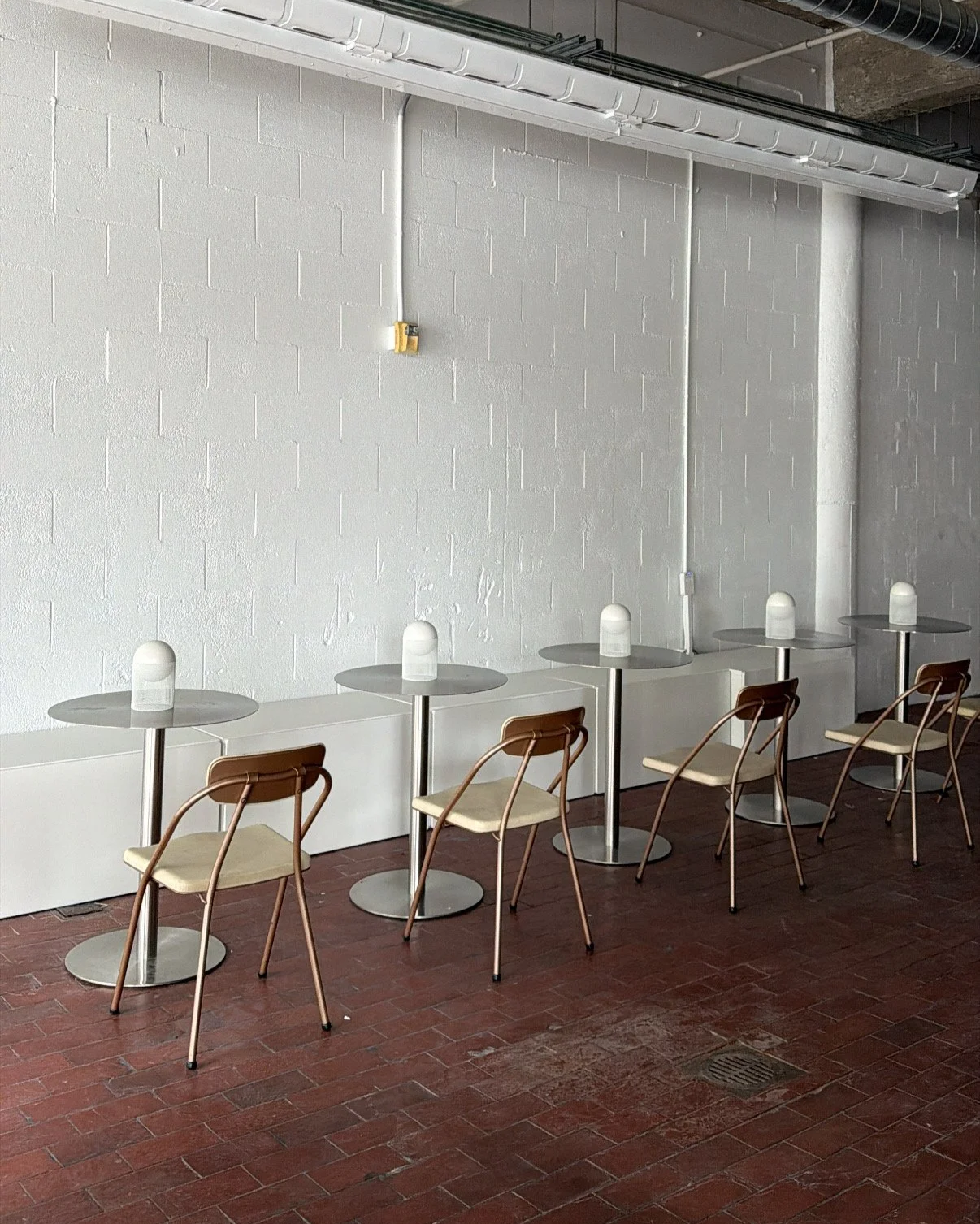Four small round tables with beige chairs, each table has a white lamp, set against a white brick wall in an industrial-style space with red brick flooring.