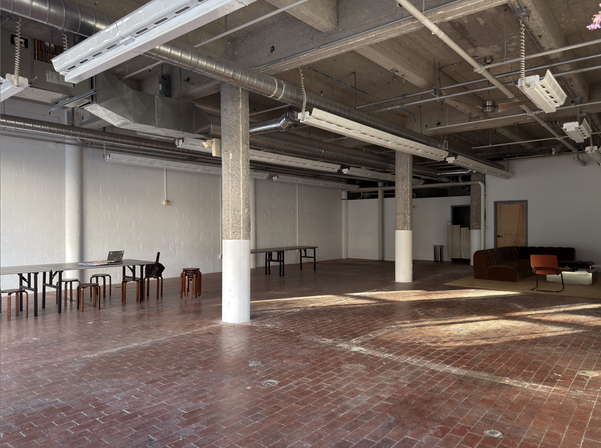 Empty industrial-style open space with brick flooring, exposed ceiling pipes, and minimal furniture including a few chairs, a table with a laptop, and a vintage sofa with an orange chair.