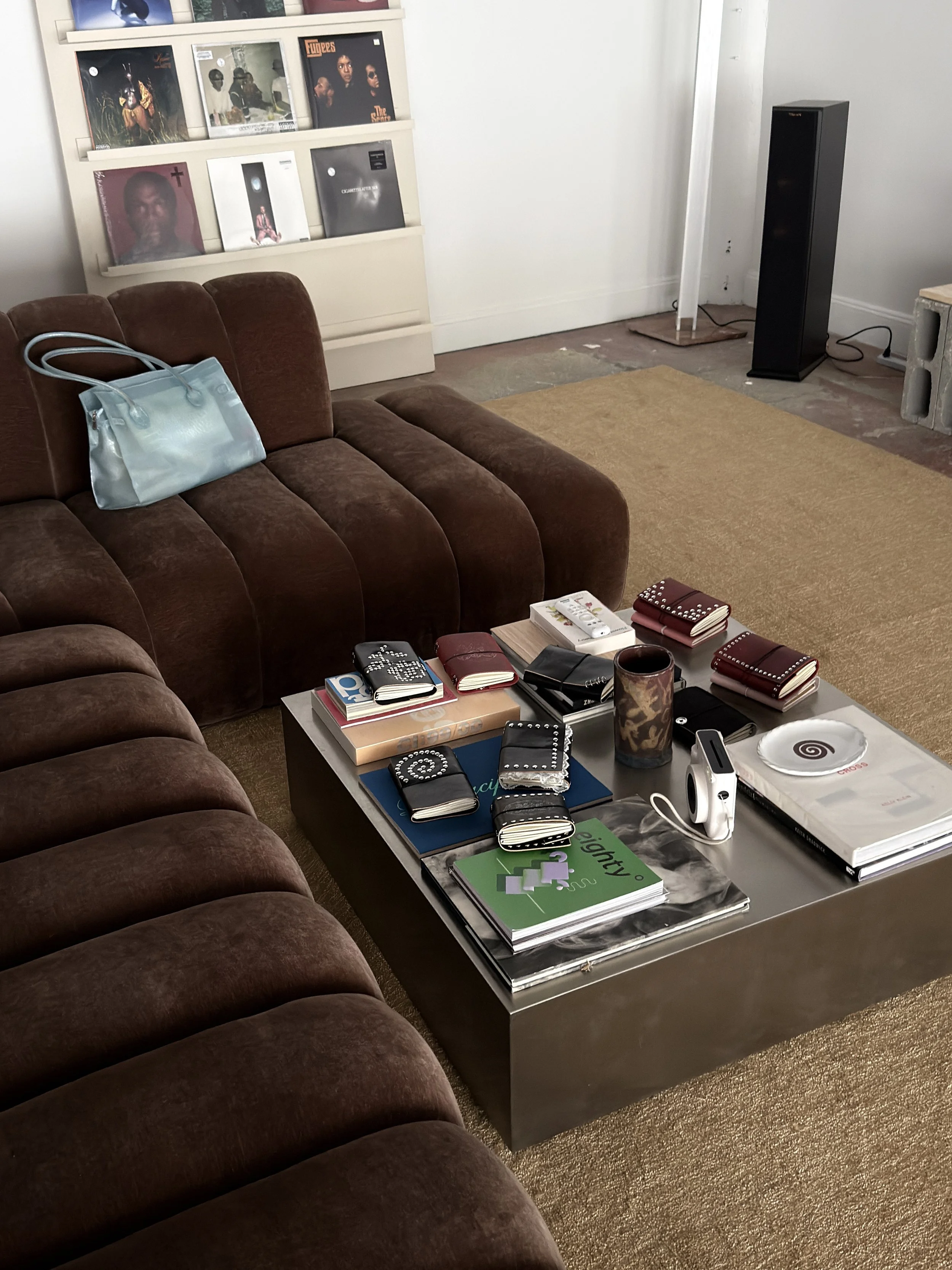 A living room with a brown velvet couch, a marble coffee table filled with notebooks, books, and decorative objects, and a wooden shelf with vinyl records against a white wall.