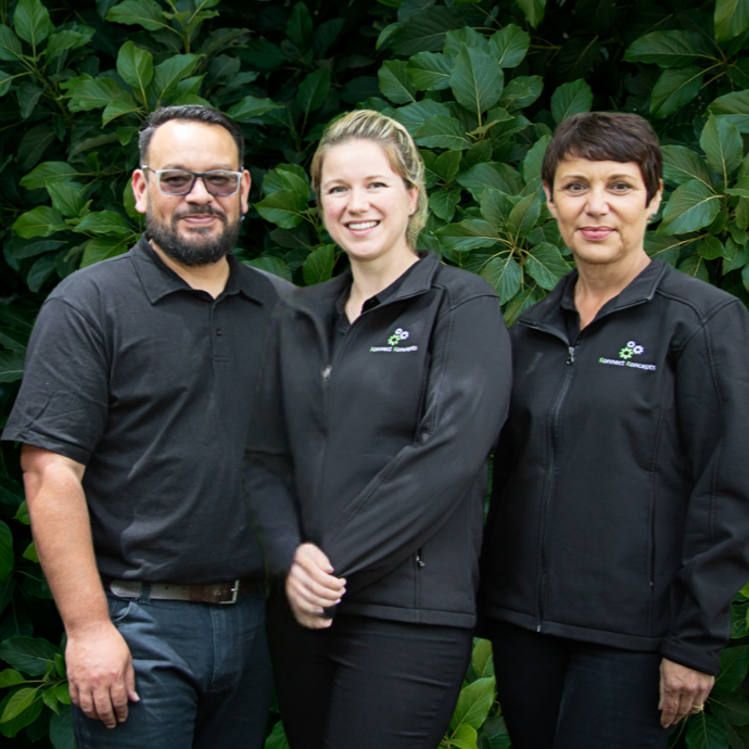 Three smiling people standing outdoors in front of large green leaves, two women and one man, dressed in black jackets or shirts.