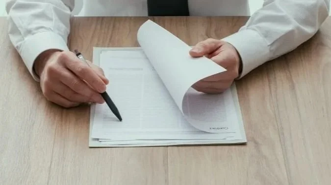 A person in a white shirt is sitting at a wooden table, using a black pen to write on or review documents.