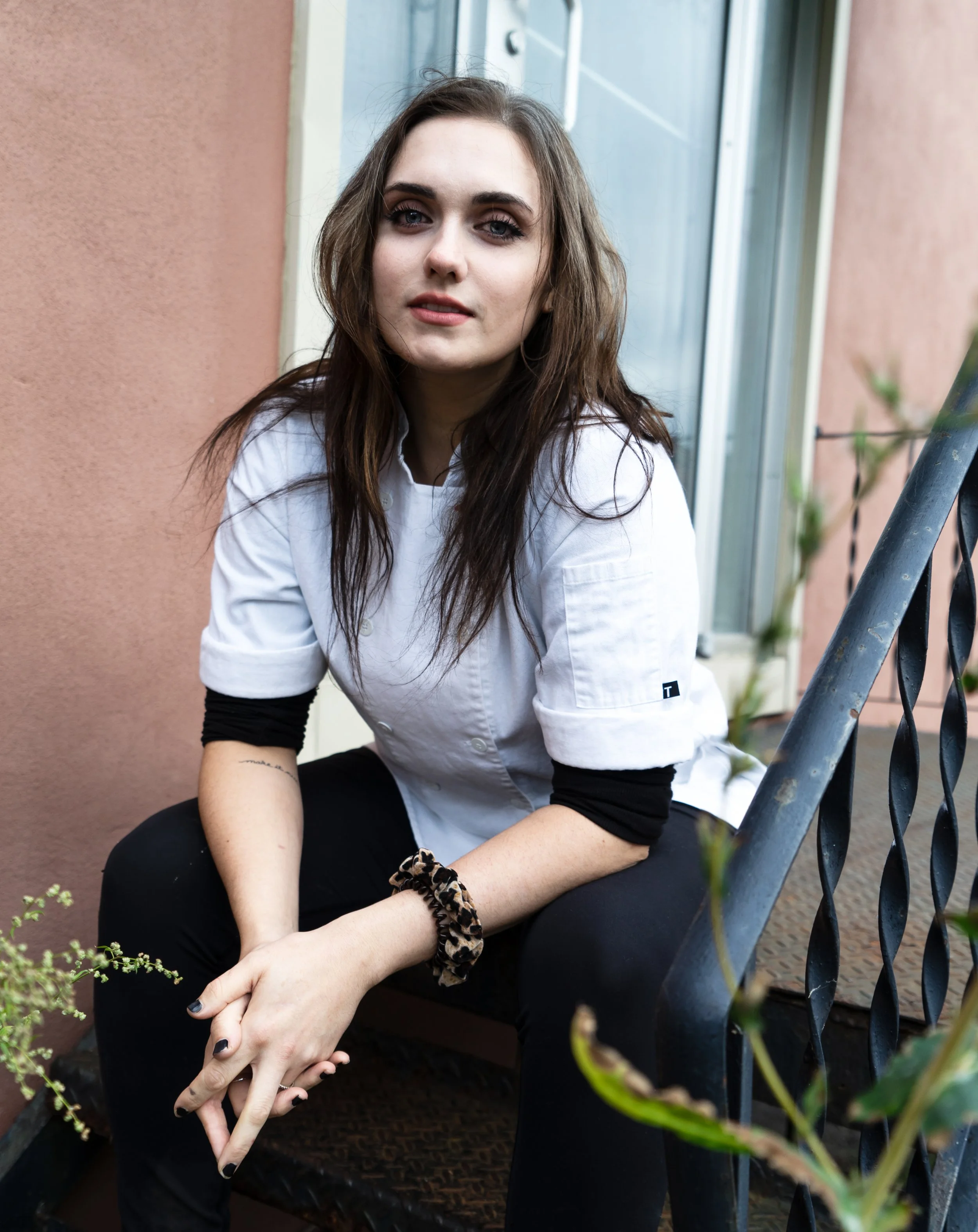 A young woman with long brown hair sitting outdoors near a pink wall, wearing a white shirt and black pants, resting her arms on her knees, with greenery and a metal railing nearby.