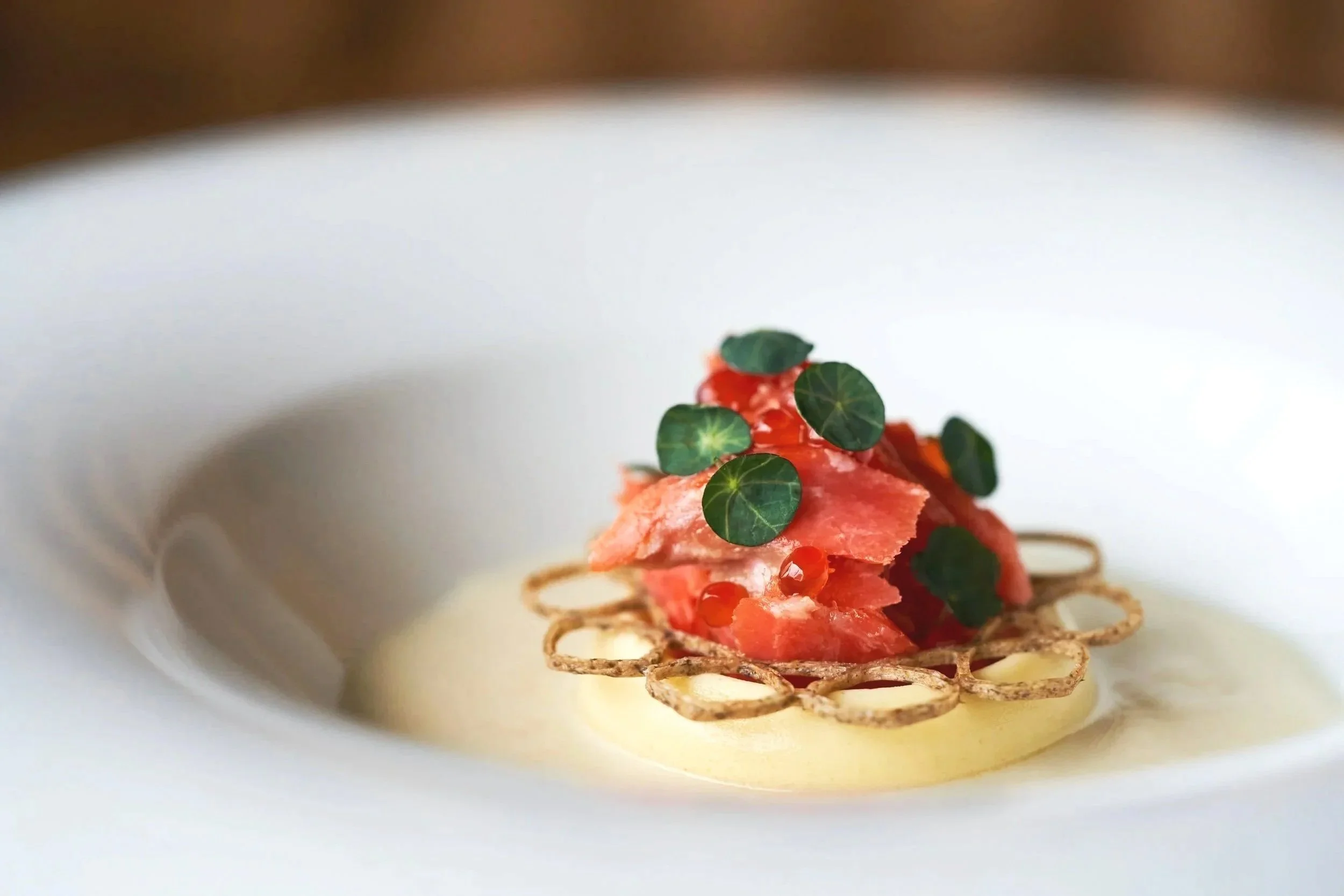 Close-up of an dish by Justin Champagne-Lagarde of Perch with thin crispy base, topped with slices of pink smoked salmon, small green leaves, and red caviar, presented on a white plate. Photographed by Gumption Studio, Ottawa food photographer. 