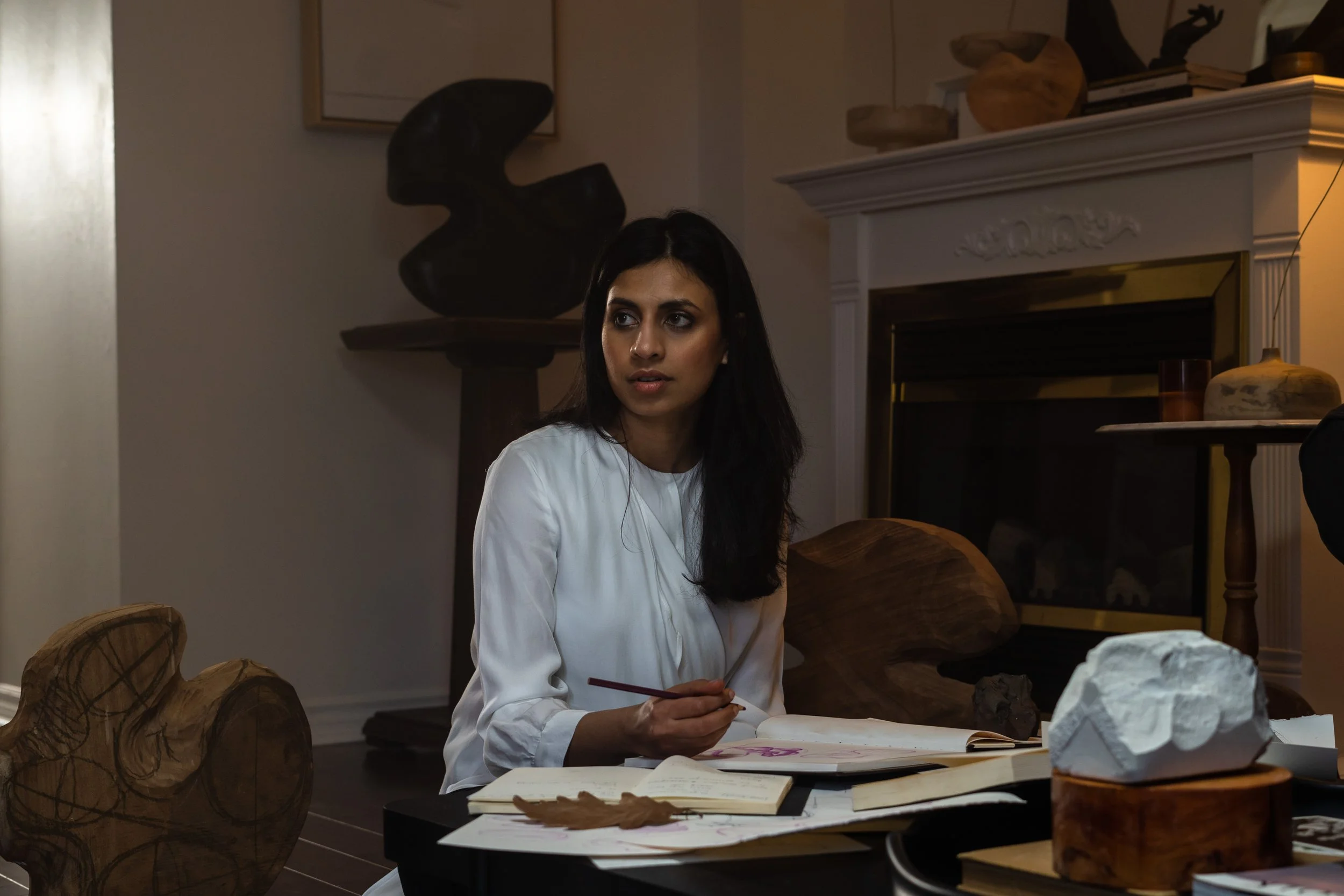A woman with long black hair wearing a white shirt sitting at a table with papers, a notebook, and a pencil in her hands, in a room with decorative wooden sculptures and a fireplace.