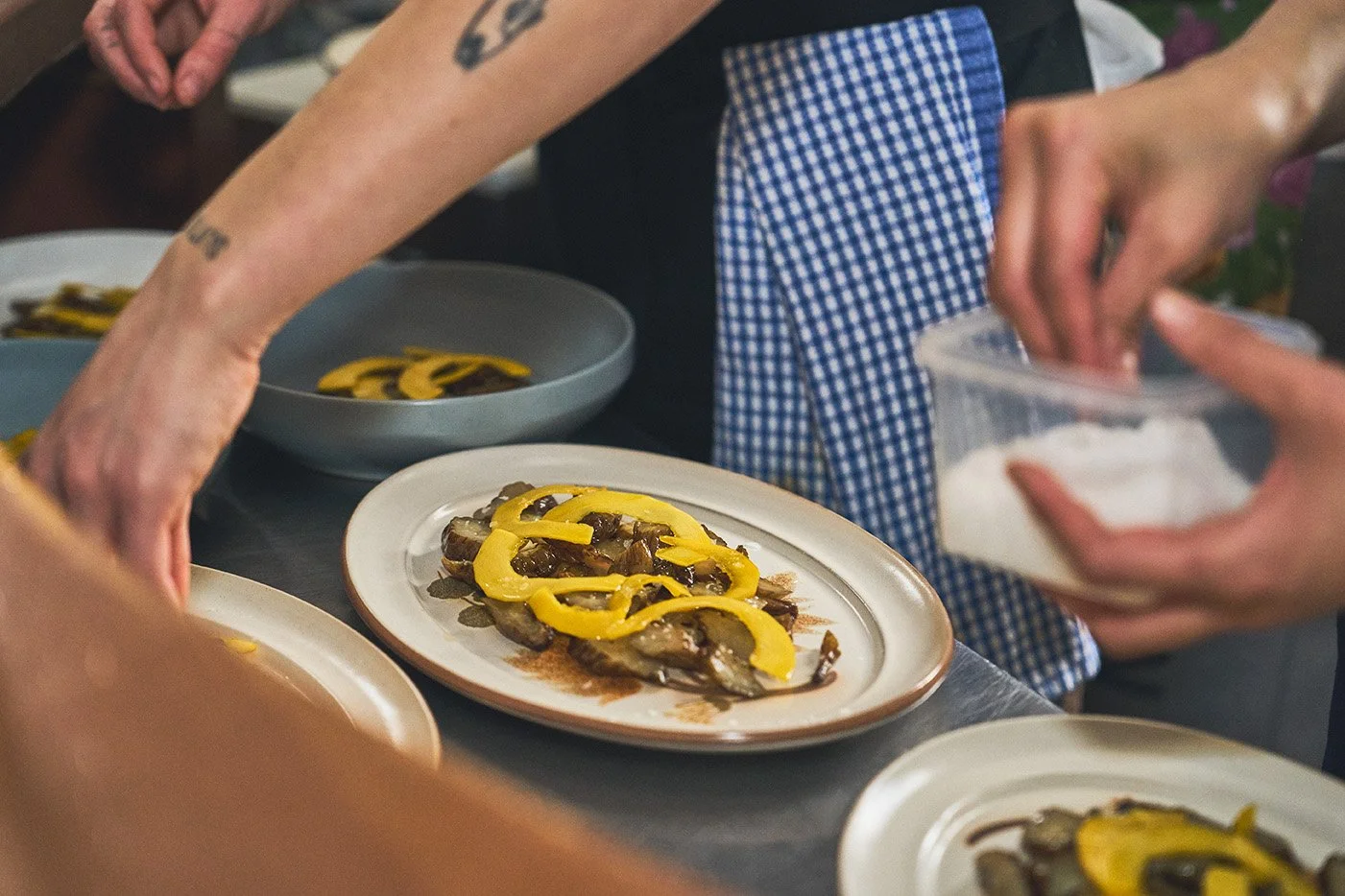 Person arranging sliced yellow bell peppers and cooked fish on a white plate at a kitchen counter, with other plates and hands visible. Photographed by Gumption Studio, Ottawa food event photographer.