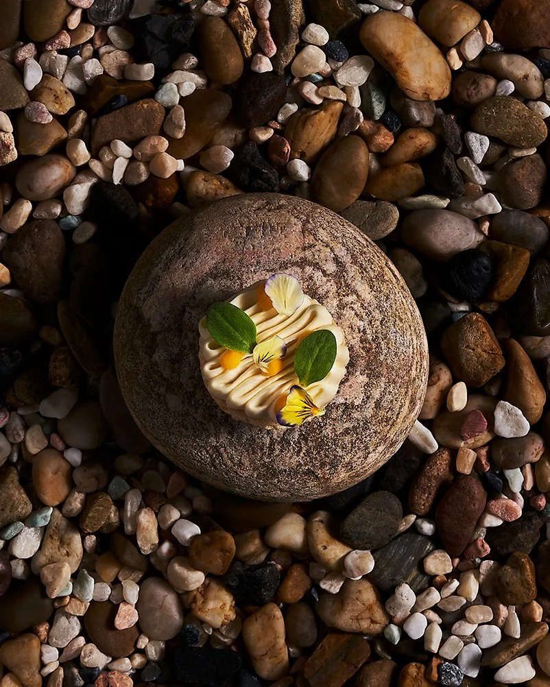Scallop macaron and edible flowers on top, surrounded by small pebbles of various colors. Photographed for the Perch Cookbook by Gumption Studio, Ottawa Cookbook photographer.