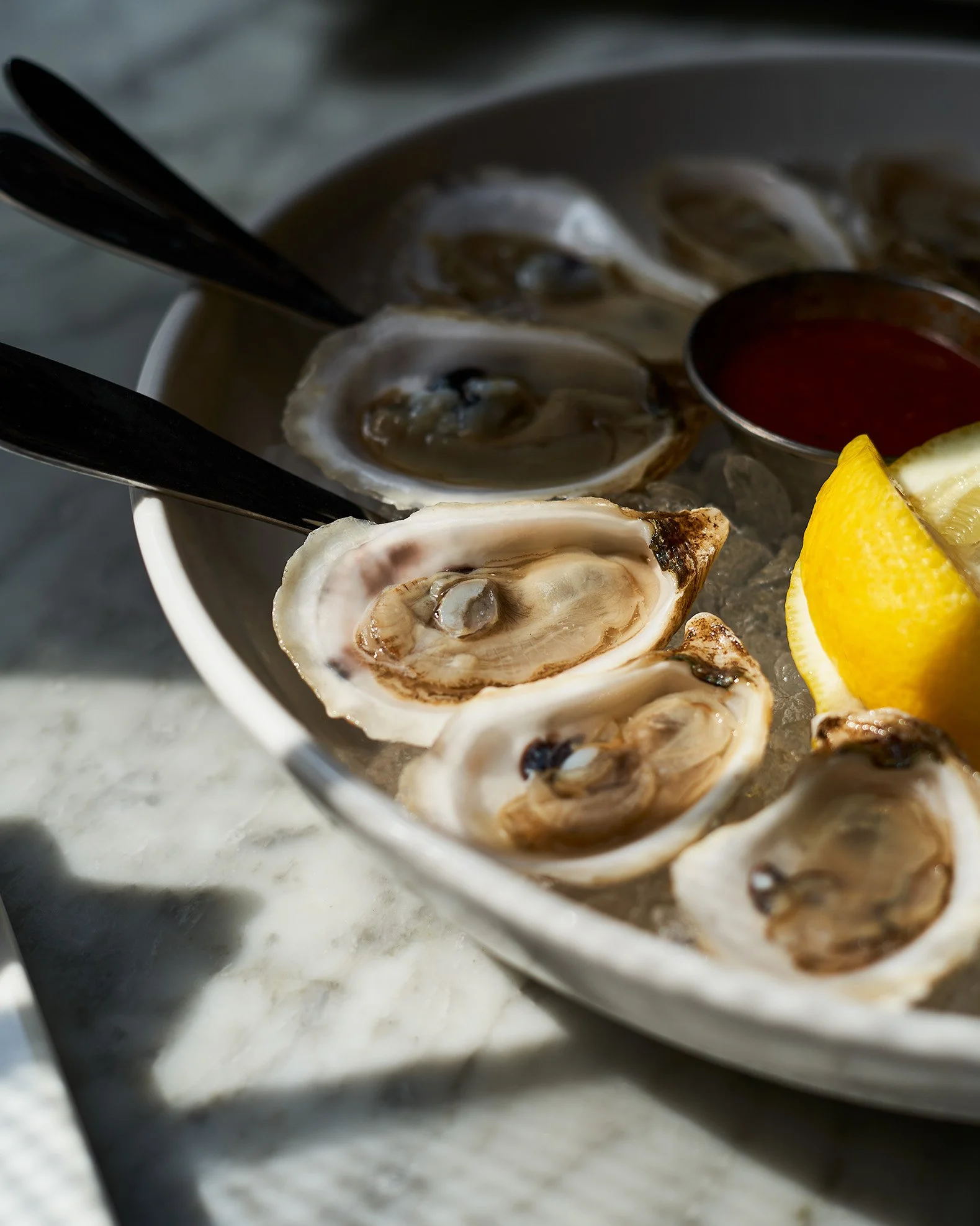 Close-up of fresh oysters at Social Restaurant with lemon wedges and cocktail sauce, with black tongs resting on it. Photographed by Gumption Studio, Ottawa food photographer.