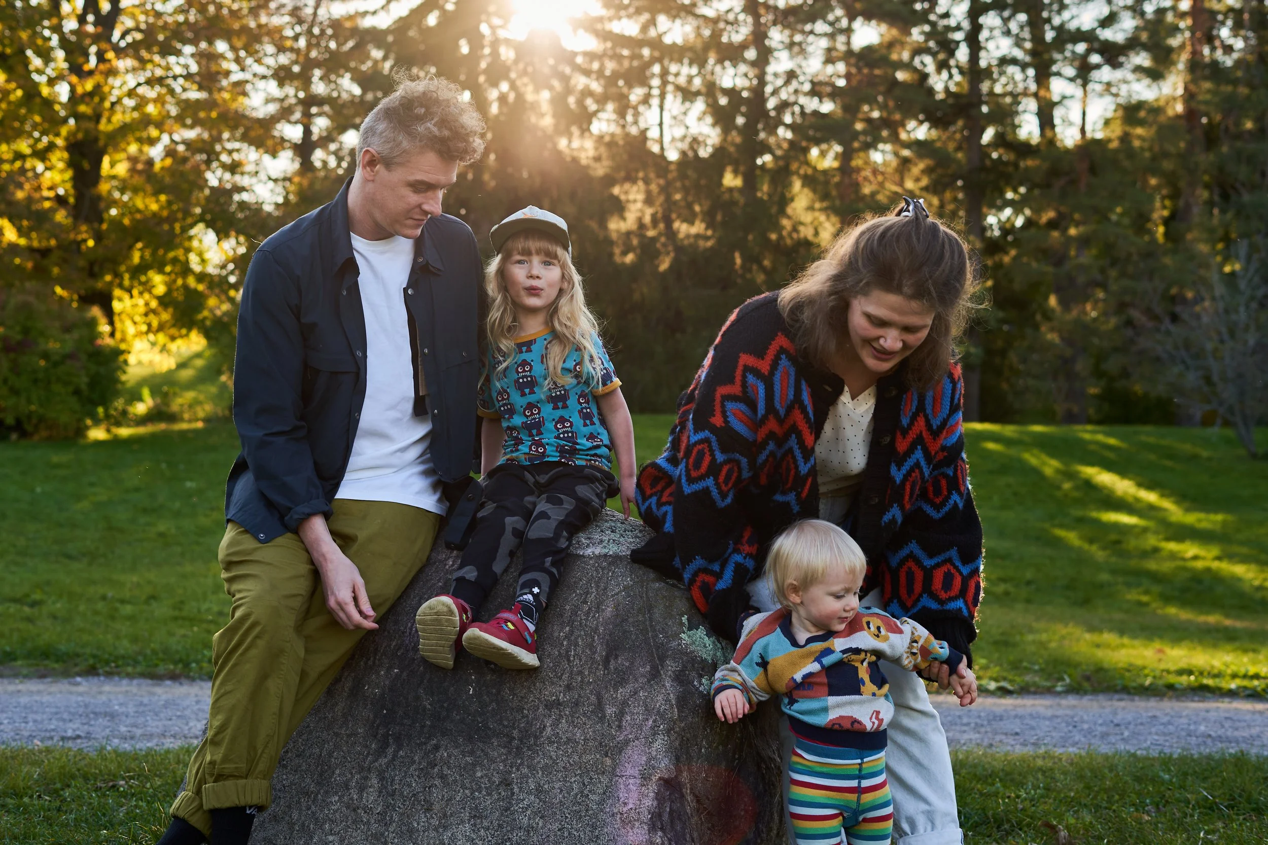 A family of four outdoors in a park during late afternoon or early evening. An adult man and woman, along with two young children, are gathered around a large fallen tree. The man has curly hair and is wearing a navy jacket and khaki pants, while the