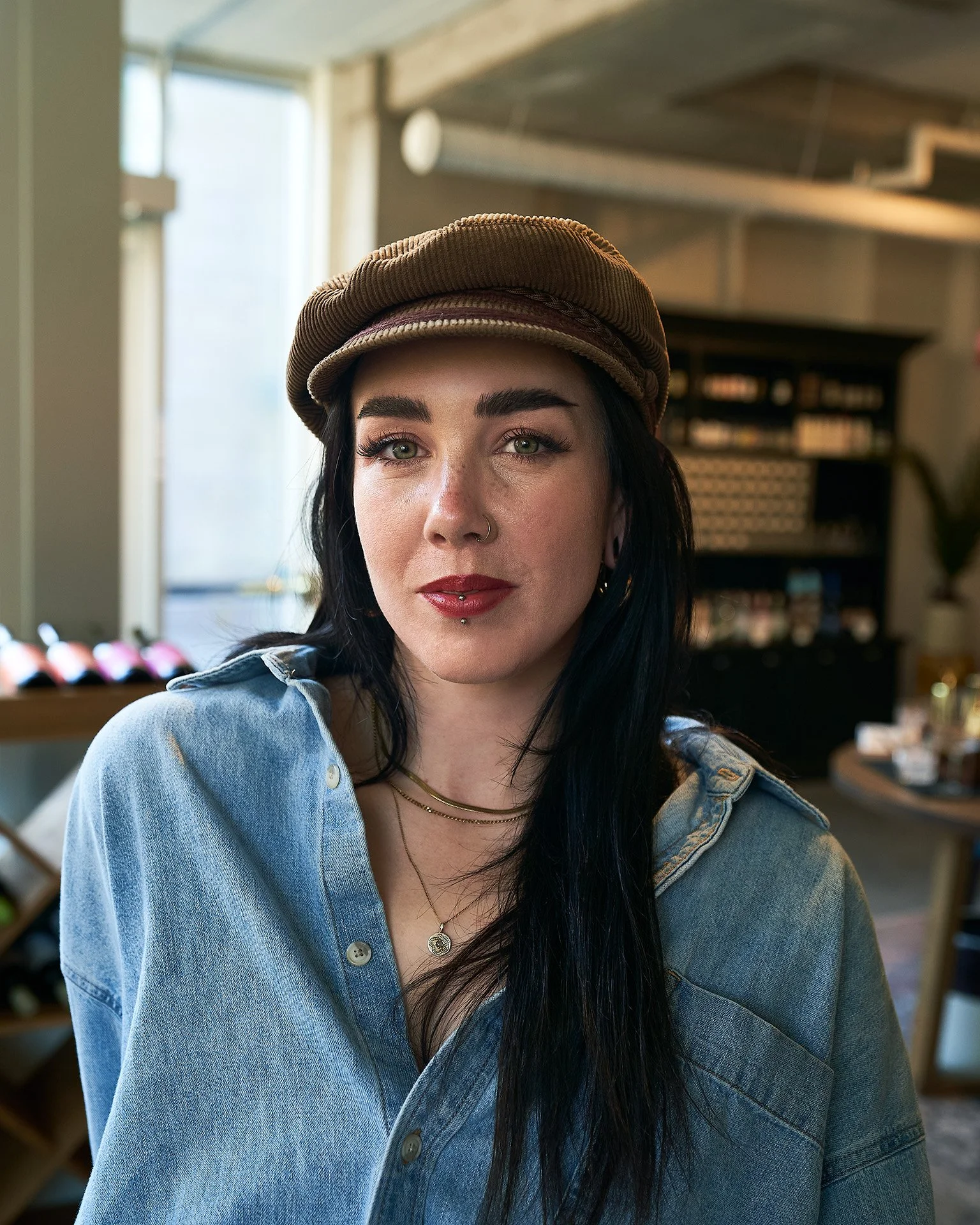 Young woman with long black hair, light makeup, nose piercings, and a silver necklace, wearing a brown hat and a denim jacket in a well-lit cafe or restaurant.