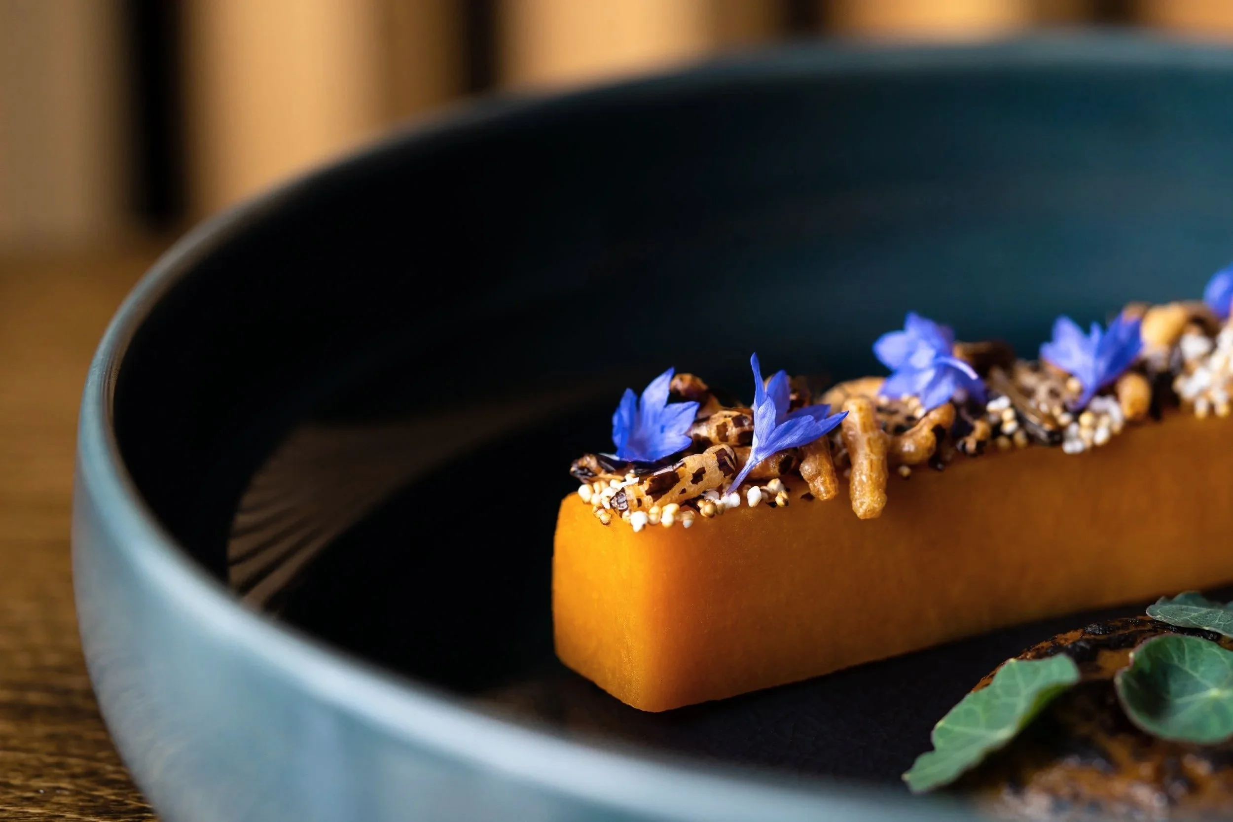 A slice of roasted butternut squash garnished with edible blue flowers, sprinkled with seeds and herbs, on a black plate. Dish by Justin Champagne of Perch. Photographed by Gumption Studio, Ottawa food photographer.
