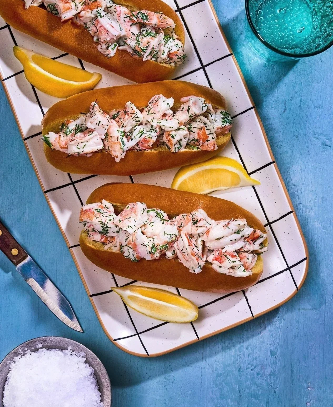 Three lobster rolls on a rectangular white plate with black grid pattern, garnished with lemon wedges, on a blue table with a glass of blue drink and a bowl of salt.