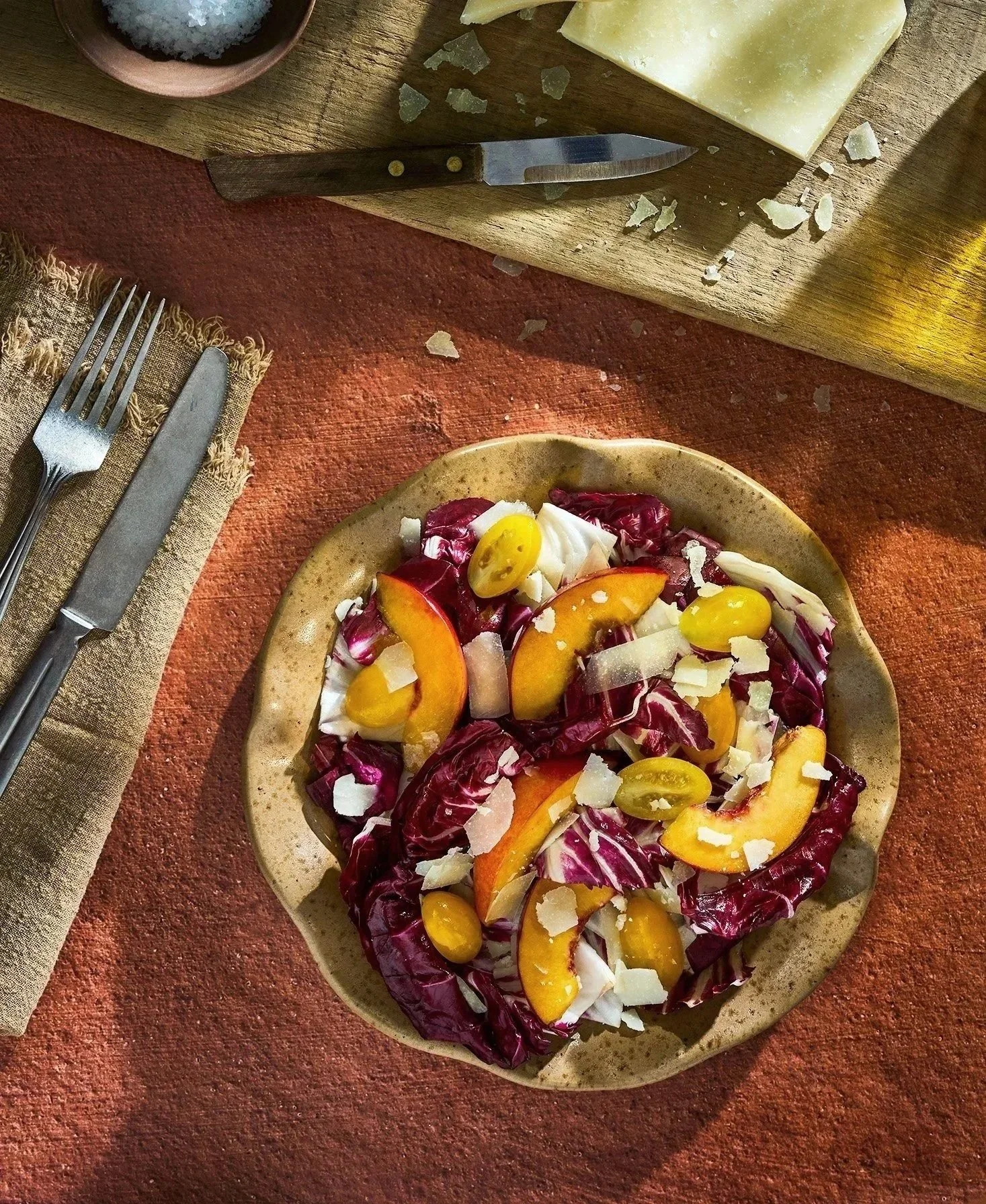 A plate of radicchio, peach slices, yellow cherry tomatoes, and shaved cheese on a rustic table with a knife on a cutting board and salt in a bowl nearby. Ingredients from Farmboy. Photographed by Gumption Studio, Ottawa commercial food photographer.