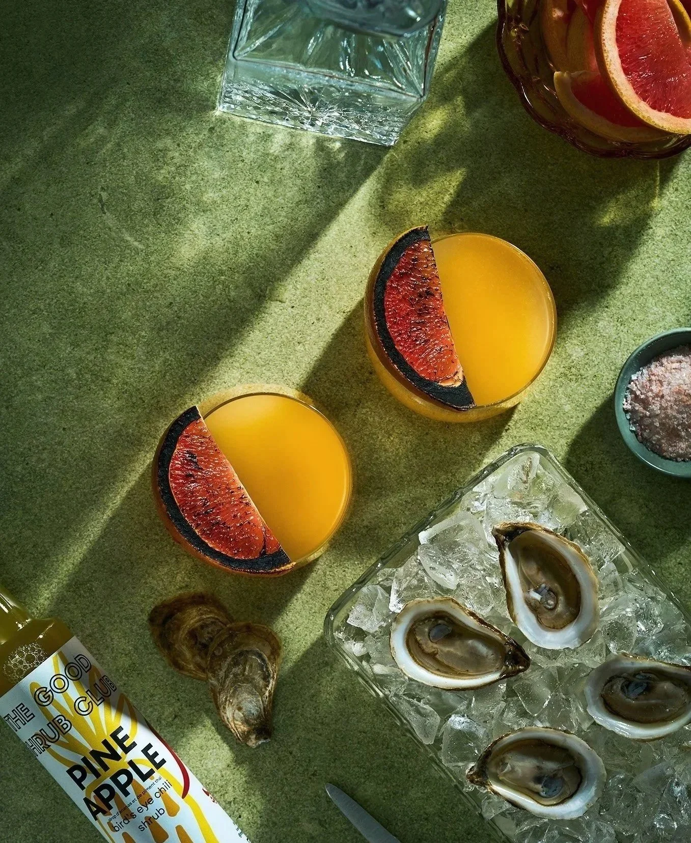 Two cocktails by Aryn Pepper of The Good Shrub Club with red grapefruit slices, oysters on ice,  on a green surface with shadows. Photographed by Gumption Studio, Ottawa commercial photographer.