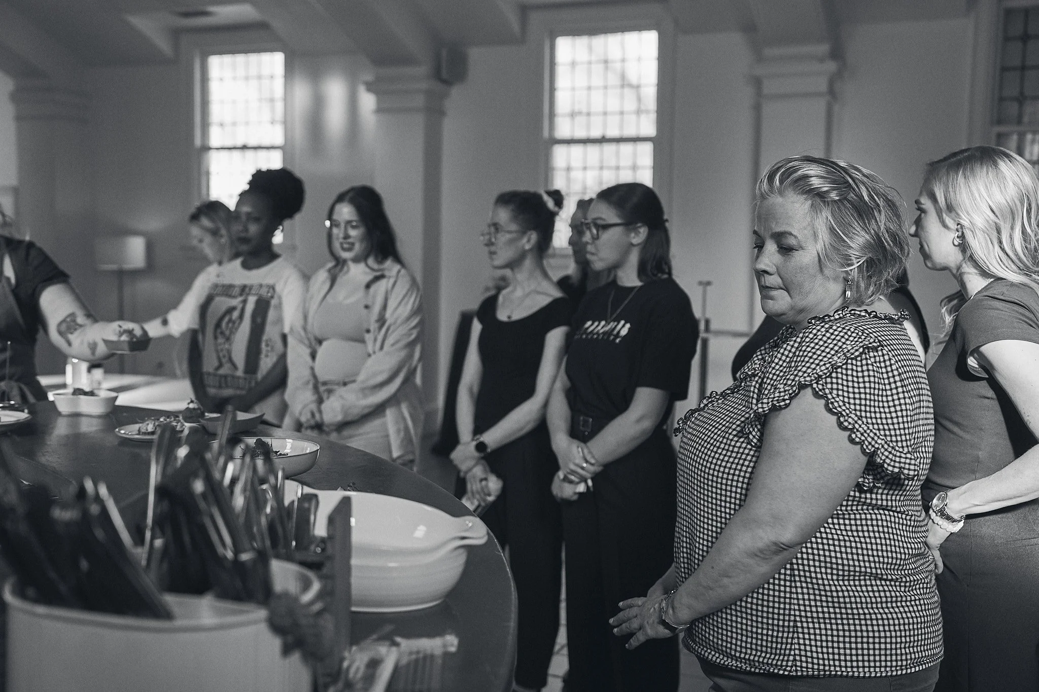 A group of women standing in a line at a table in a bright room with large windows, participating in a cooking or baking class.