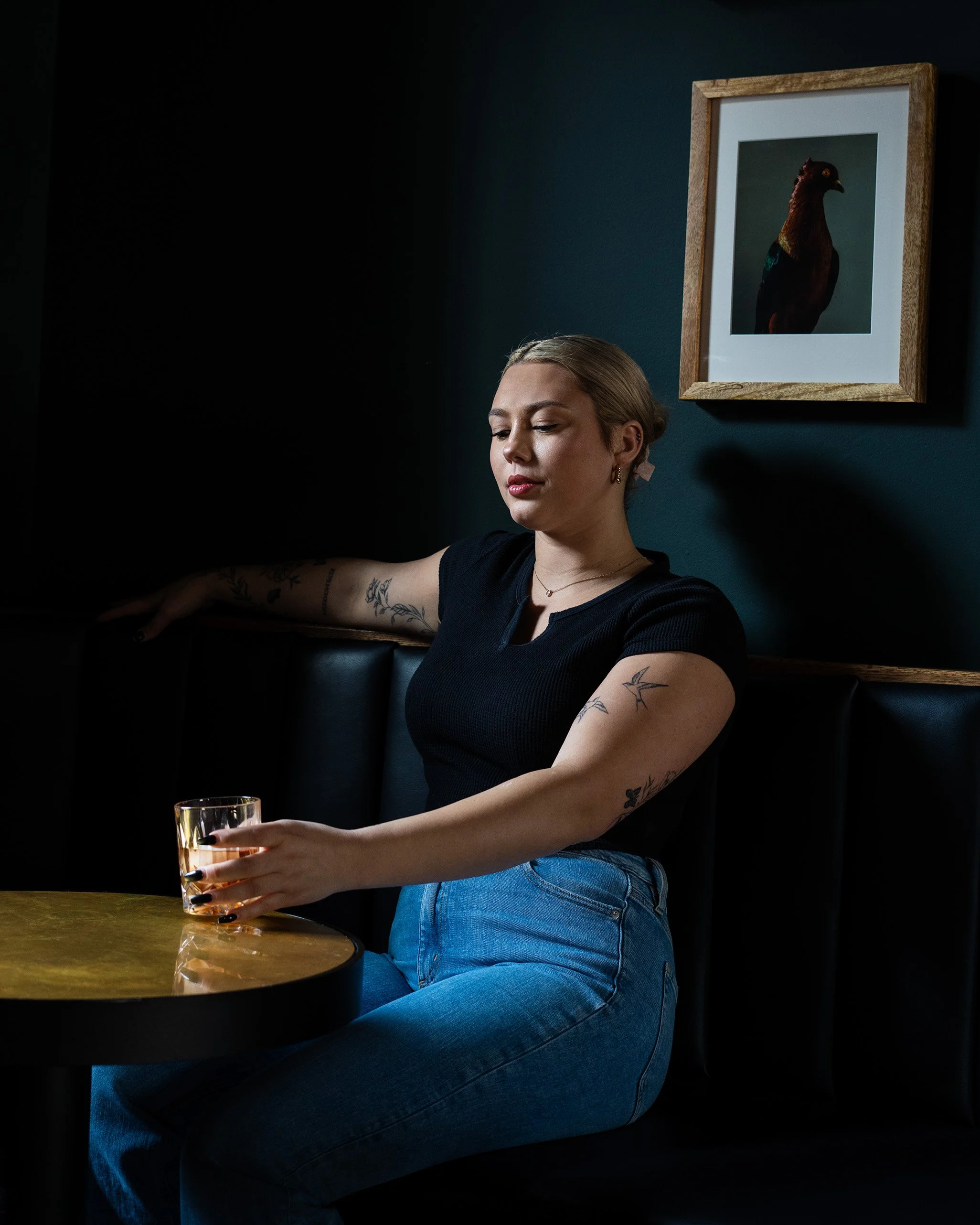A woman with tattoos on her arm sitting at a bar holding a glass of drink, with a framed bird photograph on the dark blue wall behind her.