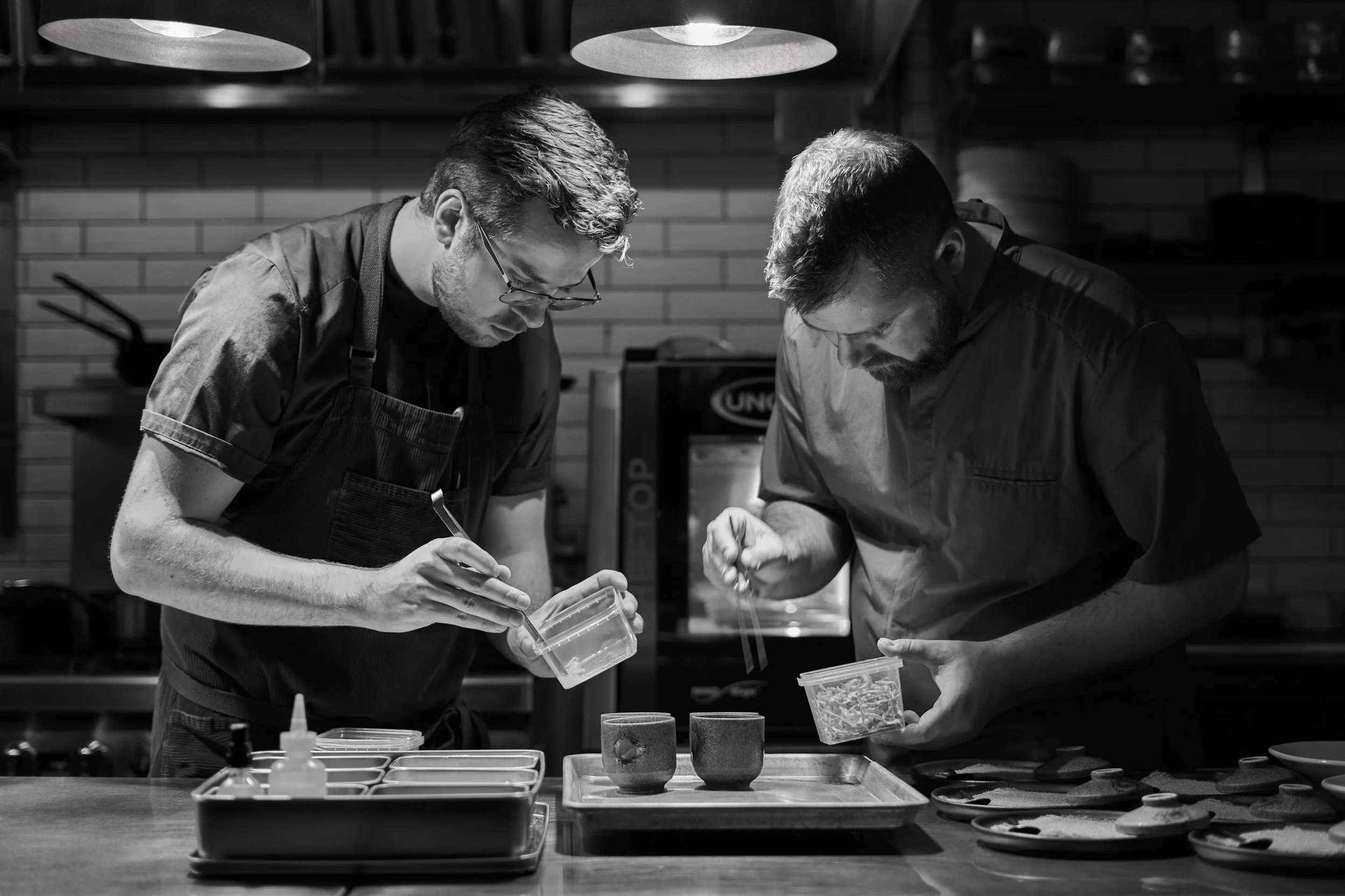 Justin Champagne preparing dishes for the Perch Cookbook. There is a brick wall background, various kitchen tools, and ingredients on the counter. Photographed by Gumption Studio, Ottawa cookbook photographer.