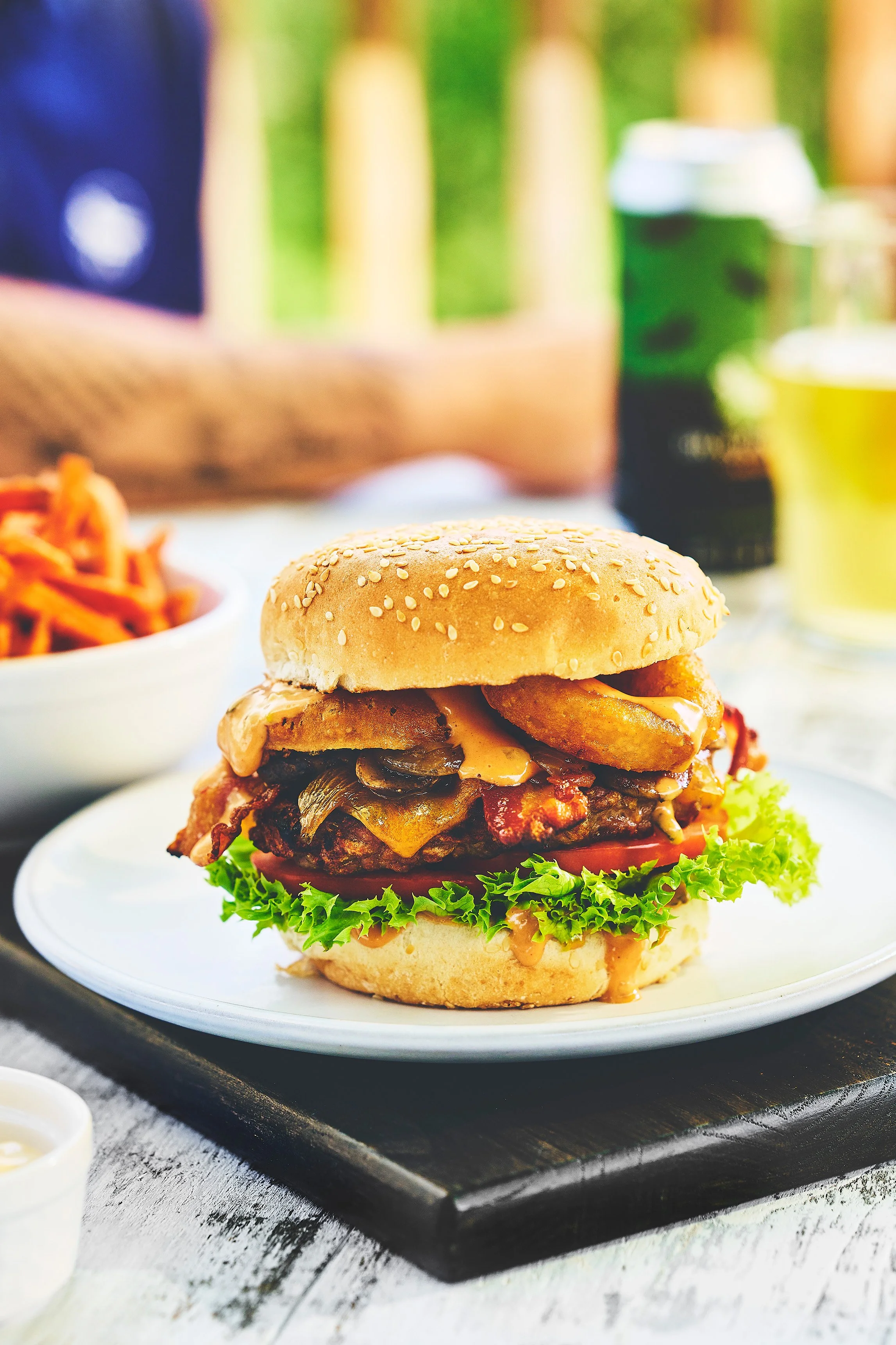 A close-up of a hamburger with lettuce, tomato, grilled mushrooms, bacon, and melted cheese in a sesame seed bun on a black tray at Dragon Fly Golf Club. Photographed by Nick Ghattas, and Ottawa food stylist Gumption Studio.