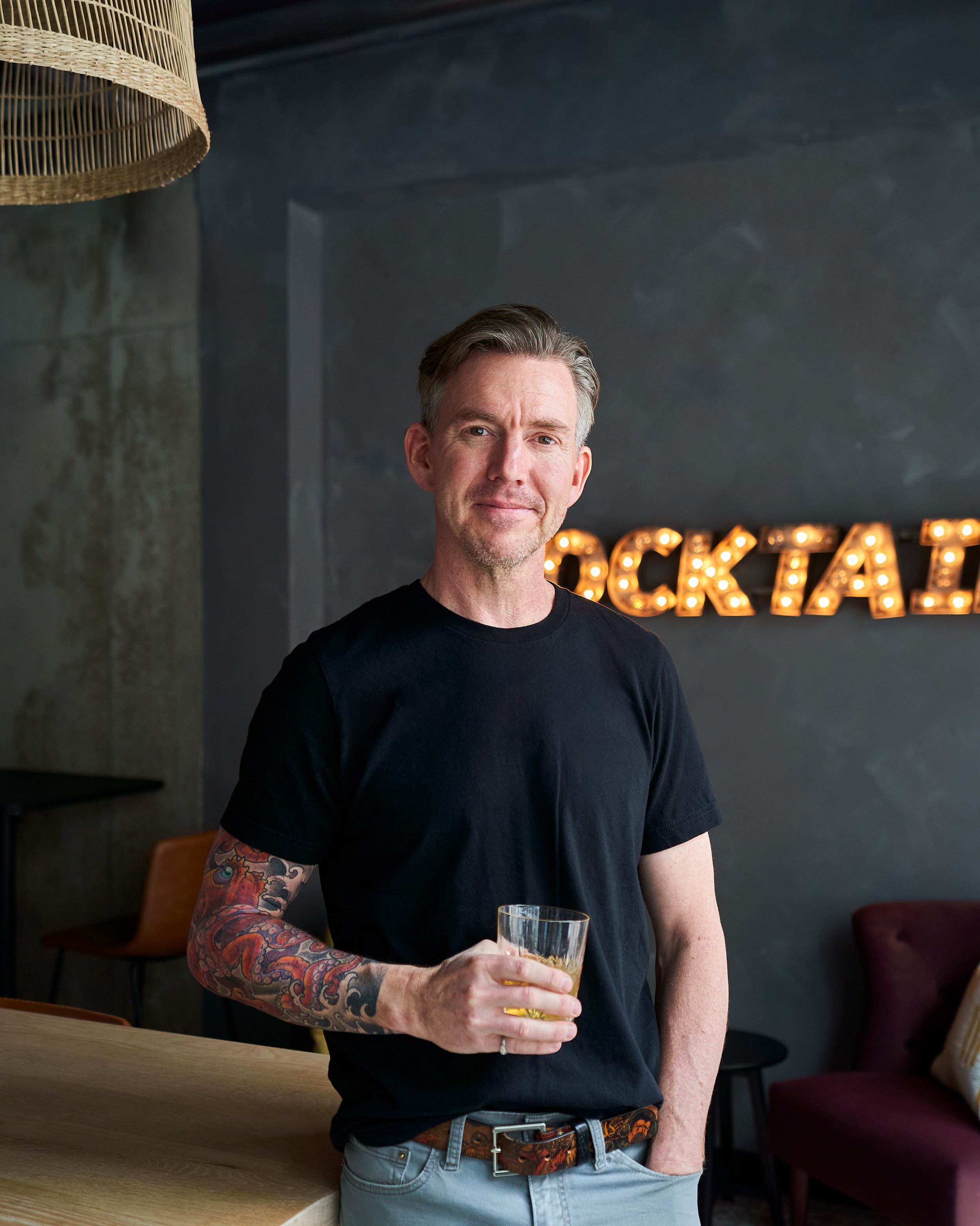 A man with gray hair and tattoos on his arm, wearing a black t-shirt and holding a glass of drink, standing in a dimly lit room with a dark wall and illuminated sign that spells 'ECKA' in the background.