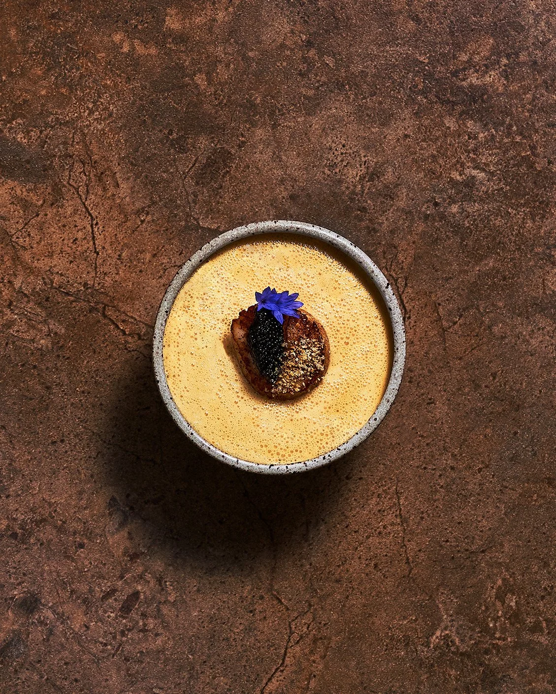 A top-down view of a scallop and caviar dish topped with a purple edible flower and black caviar, placed on a textured brown surface. Photographed for the Perch Cookbook by Gumption Studio, Ottawa Cookbook photographer.