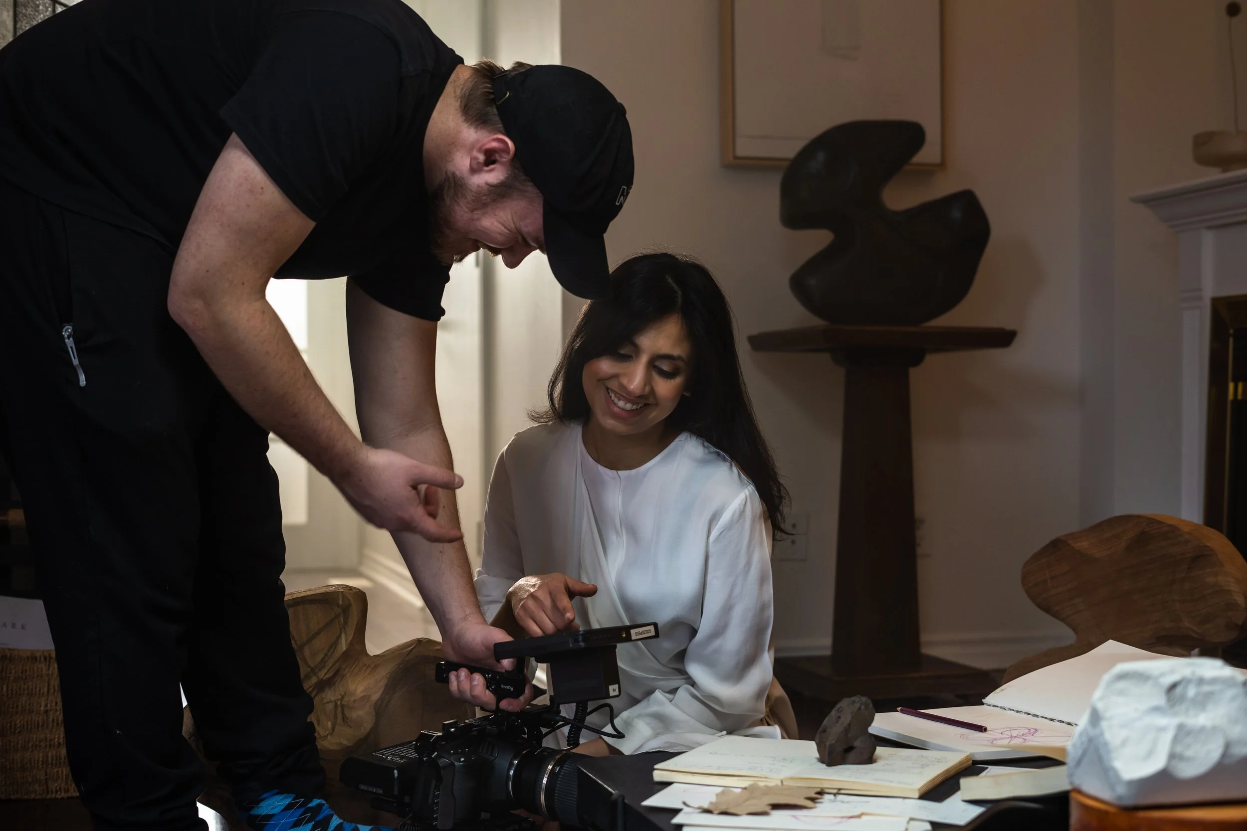 A man and woman laughing and looking at a camera or video monitor on a table surrounded by papers and notebooks in a room with artwork on the walls.