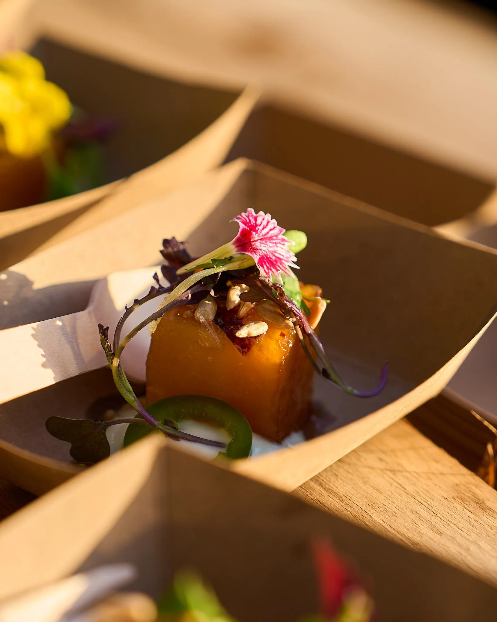 Close-up of a dish from Harvest: A Feast of Fall with pink petals, green stem, and delicate purple leaves, placed on a wooden surface inside a paper takeout container. Photographed by Gumption Studio, Ottawa event photographer.