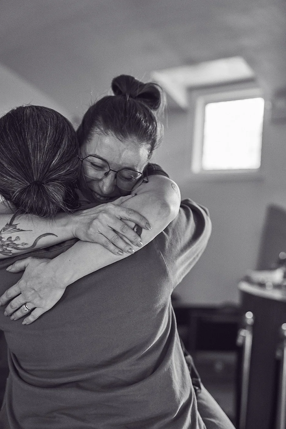 Two women hugging, one with glasses and the other with tattoos, in a room with a window and blurred furniture in the background at the Girl Dinner Event at Parlour. Photographed by Gumption Studio, Ottawa event photographer.