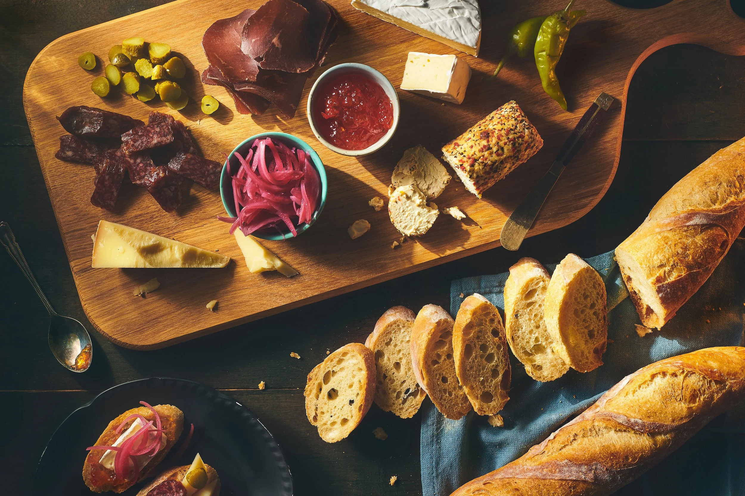 A charcuterie board with cheeses, meats, pickles, olives, Your Bread Box baguette on a dark table. Photographed by Nick Ghattas, food styling by Gumption Studio.