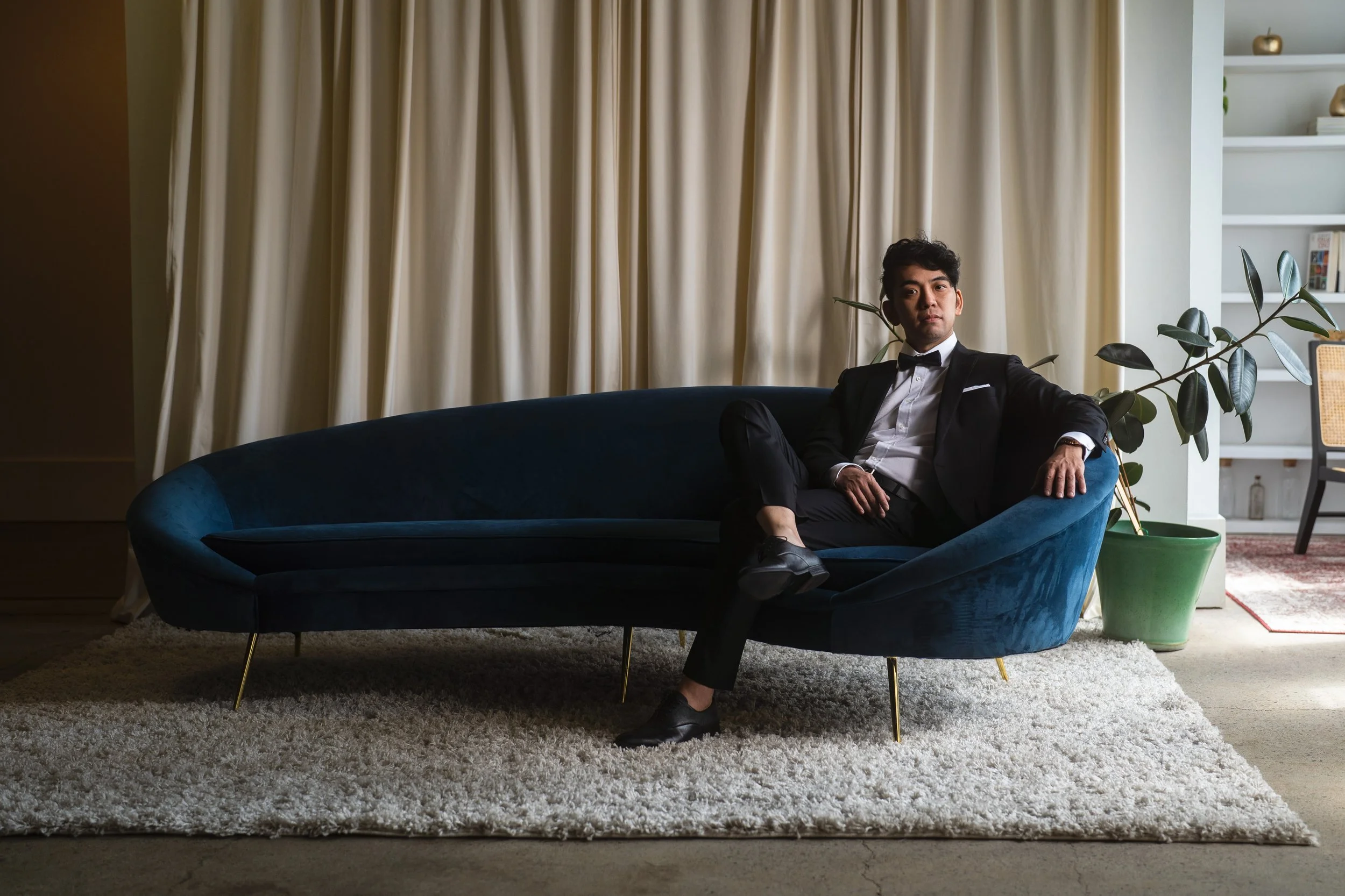 A man in a black tuxedo with a bow tie sitting on a dark blue curved sofa in a modern living room with a beige carpet, a large white curtain, a green potted plant, and white shelves with decorative items.
