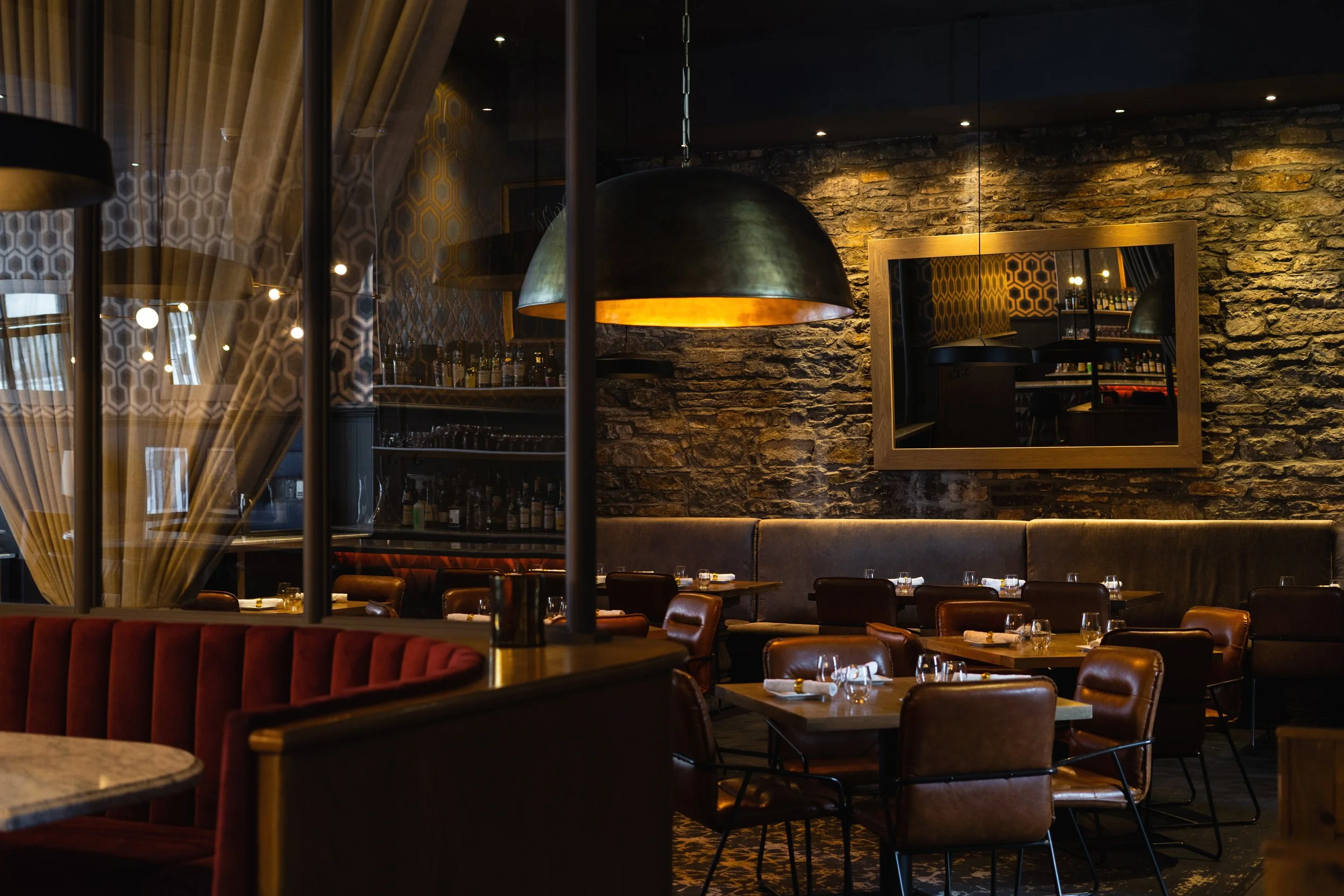 Interior of a Social Restaurant with brick walls, large black and gold pendant lights, a large mirror, tables set with glasses and napkins, and velvet chairs. Photographed by Gumption Studio, Ottawa interior photographer.