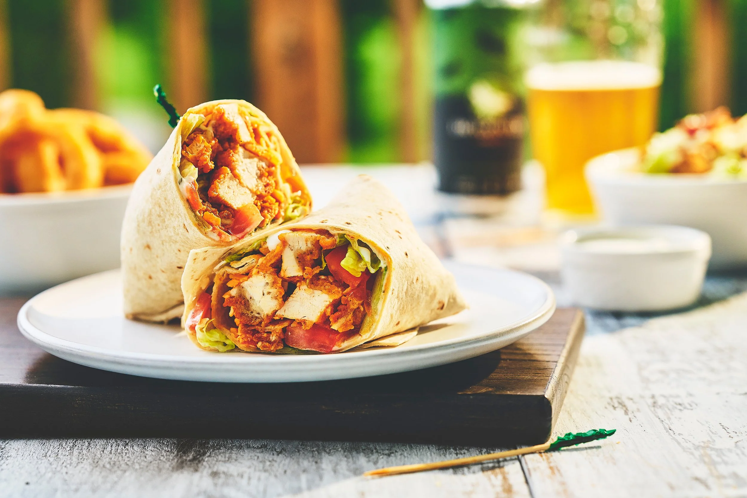 A wrap cut in half with grilled chicken, lettuce, tomato, and sauce inside, placed on a wooden table with a blurred background including a glass of beer from Dragon Fly Golf Club. Photographed by Nick Ghattas, and Ottawa food stylist Gumption Studio.